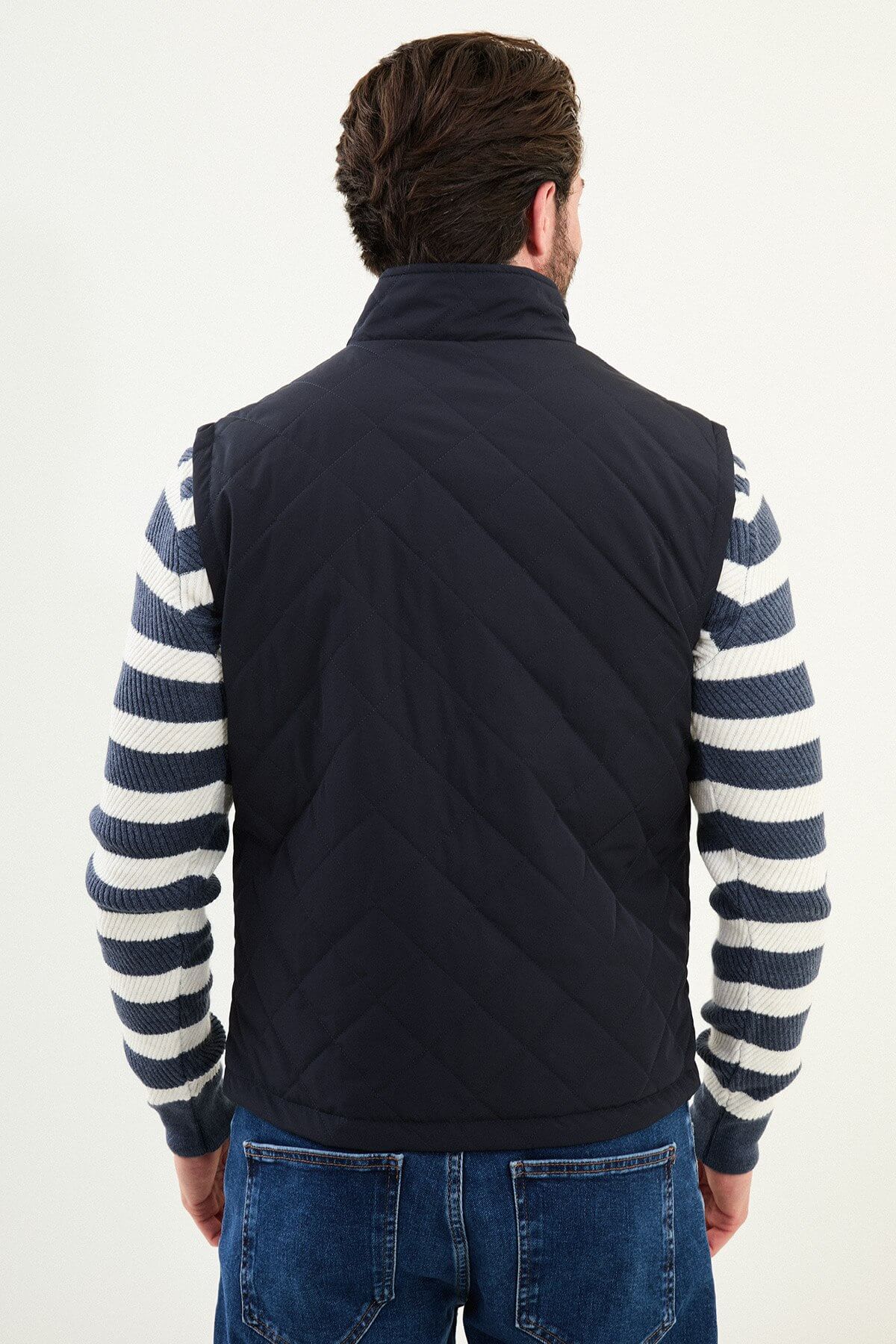 Men’s navy blue diamond-quilted technical vest with a matte finish, styled over a striped sweater for winter layering.