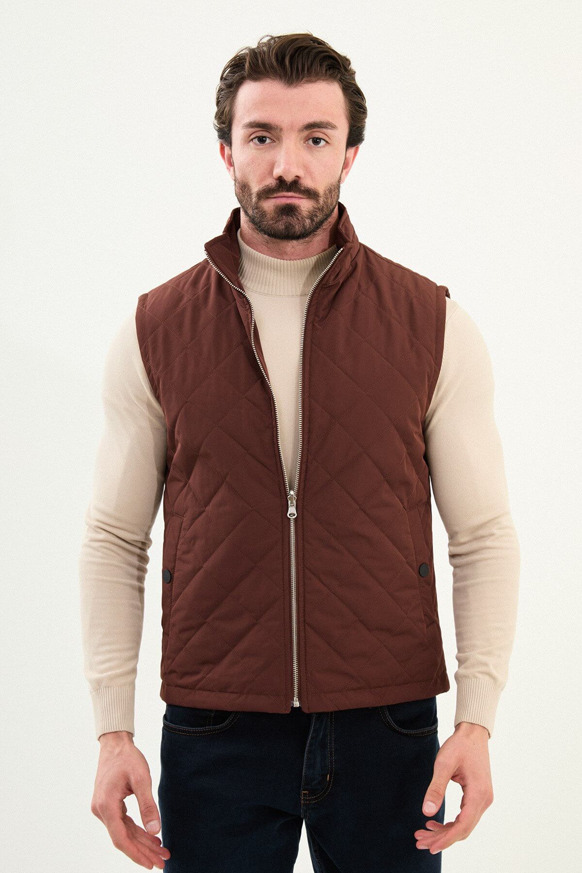 Tile rust diamond-quilted men’s technical vest in matte polyester, styled over a beige turtleneck with dark denim.