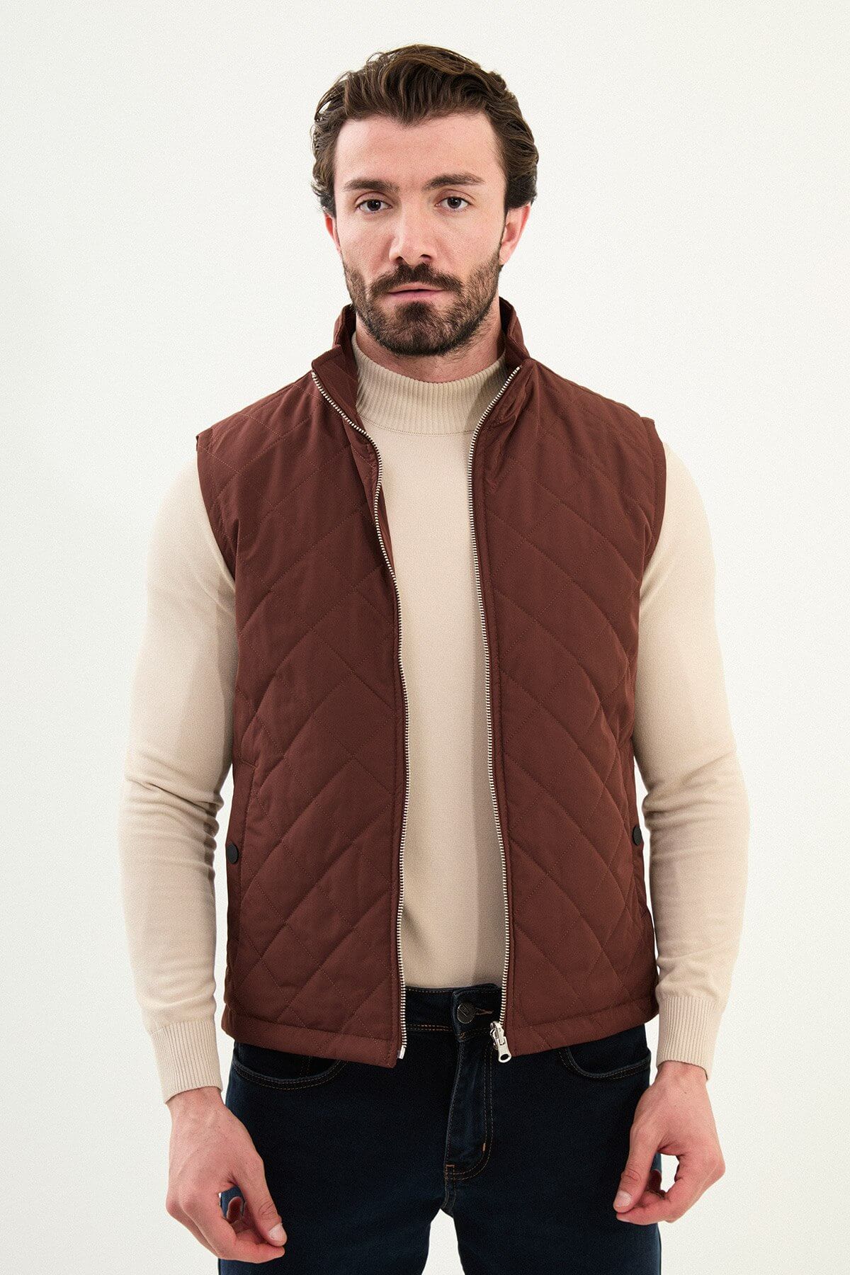 Tile rust diamond-quilted men’s technical vest in matte polyester, styled over a beige turtleneck with dark denim.