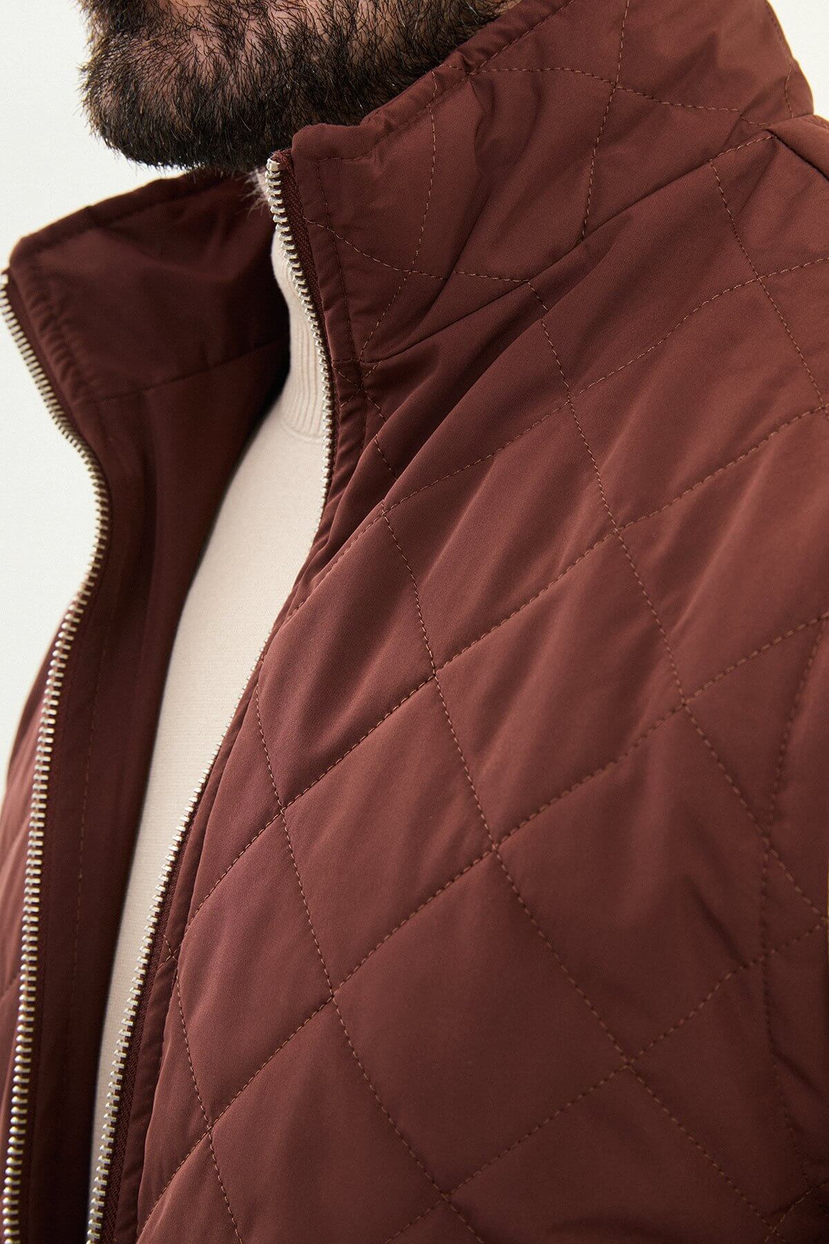 Tile rust diamond-quilted men’s technical vest in matte polyester, styled over a beige turtleneck with dark denim.