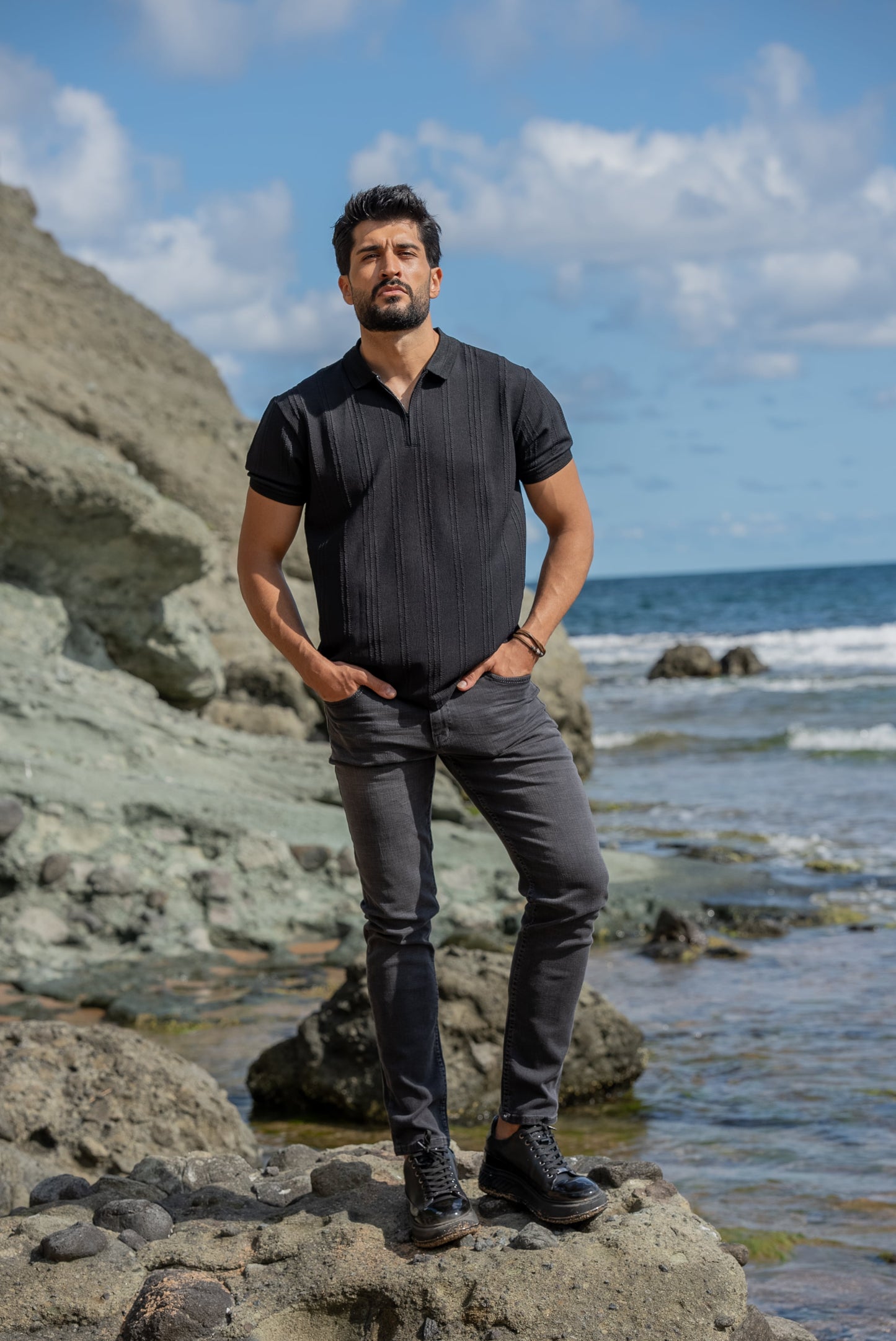 A man stands confidently on rocky terrain by the seaside, wearing a black knitted polo collar t-shirt, dark gray slim-fit jeans, and black high-top sneakers. 