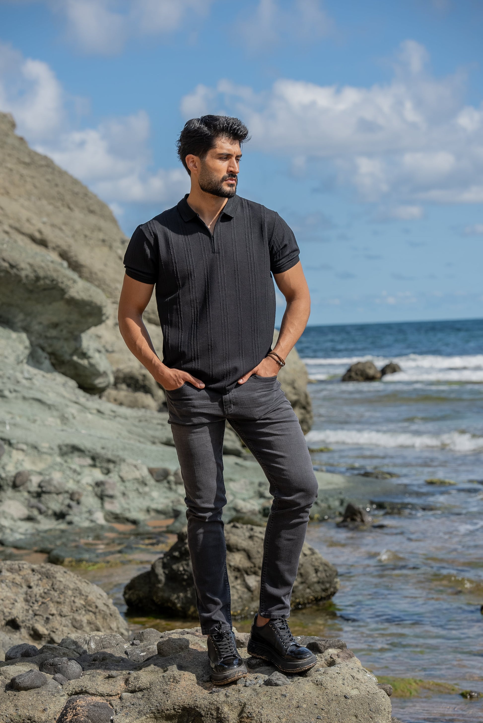A man stands confidently on rocky terrain by the seaside, wearing a black knitted polo collar t-shirt, dark gray slim-fit jeans, and black high-top sneakers. 
