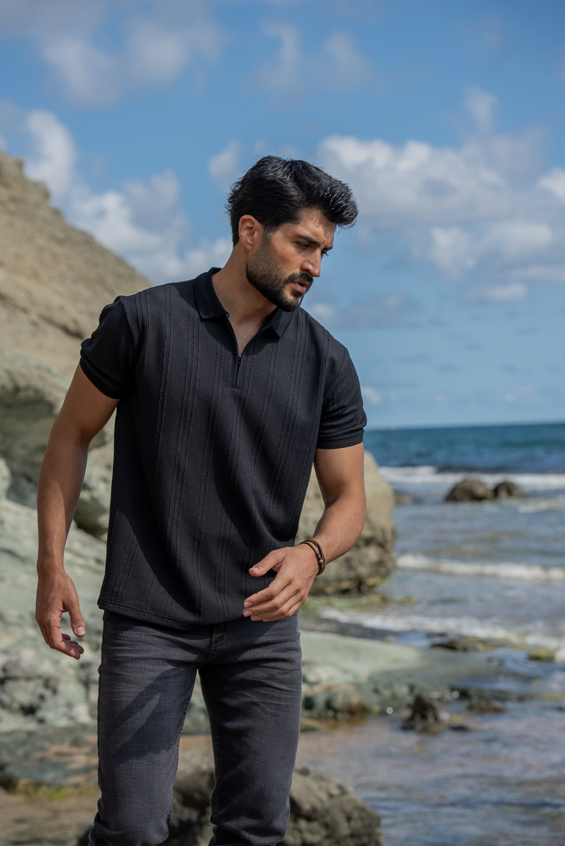 A man stands confidently on rocky terrain by the seaside, wearing a black knitted polo collar t-shirt, dark gray slim-fit jeans, and black high-top sneakers. 