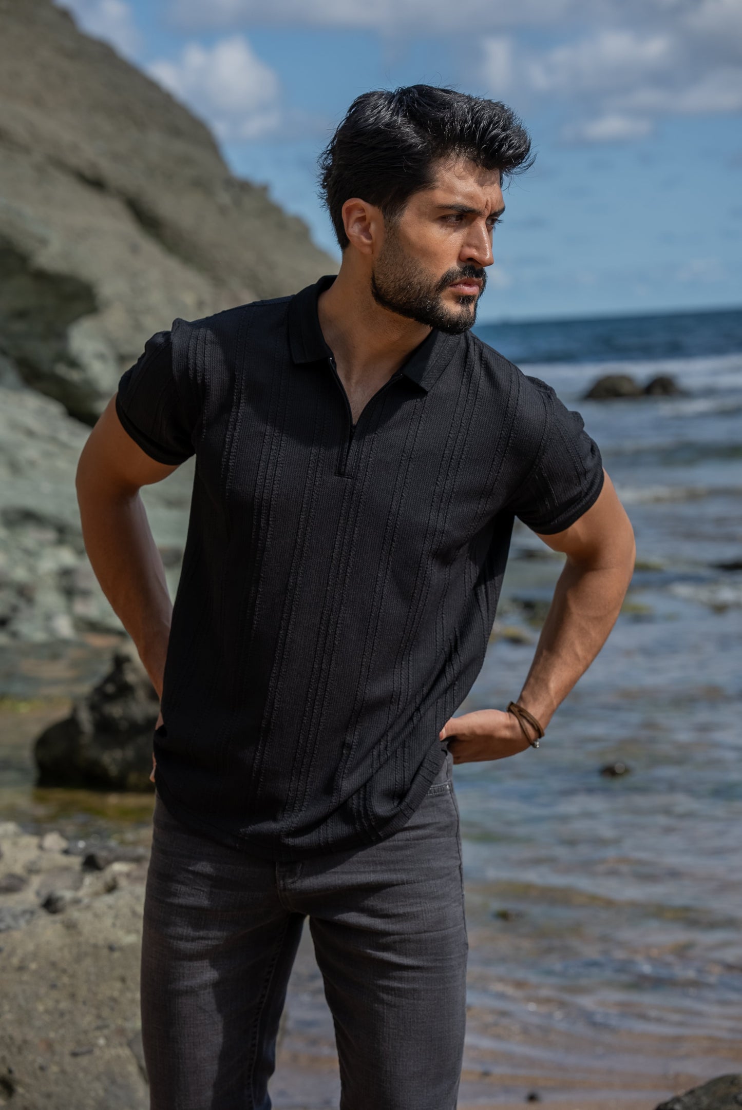 A man stands confidently on rocky terrain by the seaside, wearing a black knitted polo collar t-shirt, dark gray slim-fit jeans, and black high-top sneakers. 