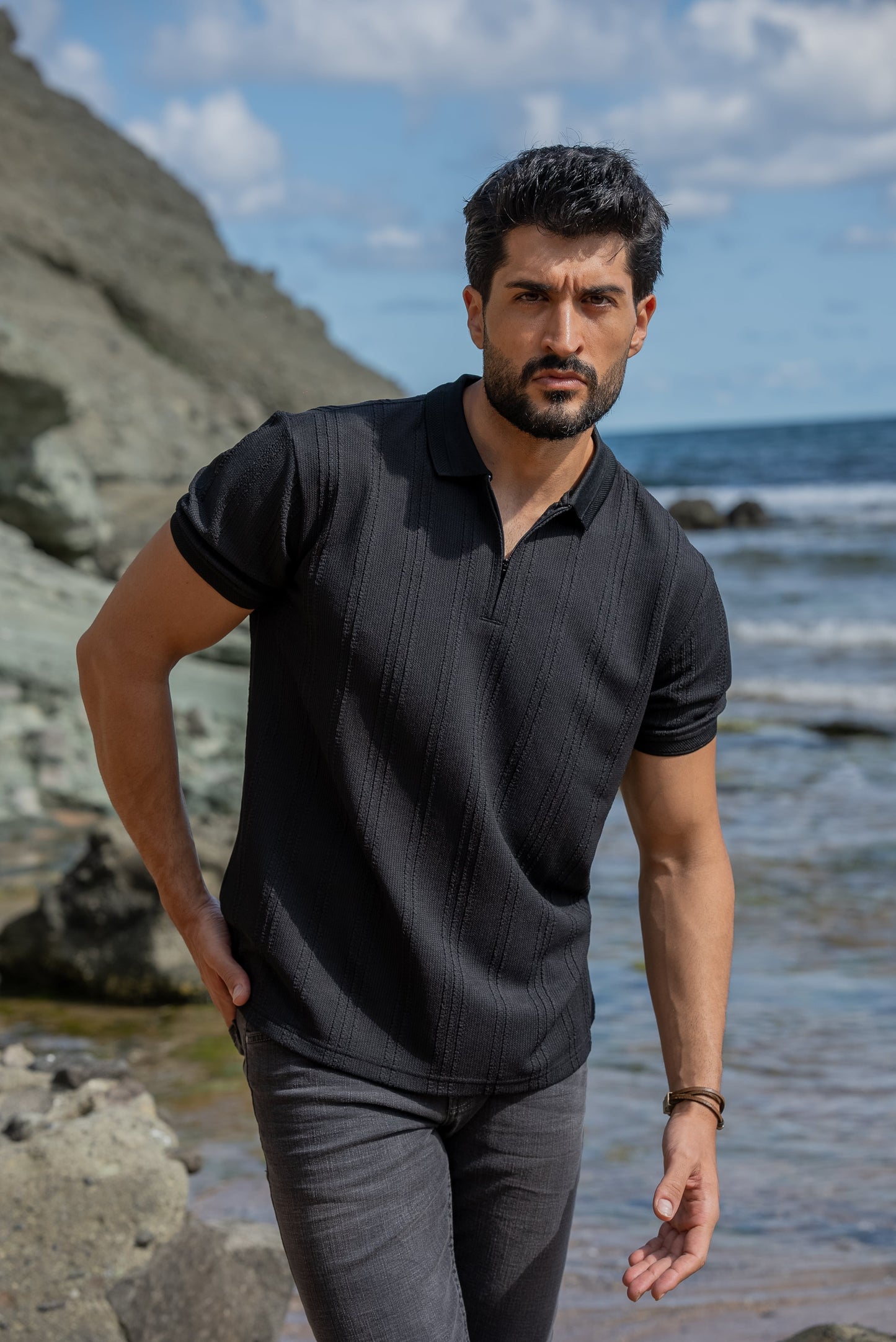 A man stands confidently on rocky terrain by the seaside, wearing a black knitted polo collar t-shirt, dark gray slim-fit jeans, and black high-top sneakers. 