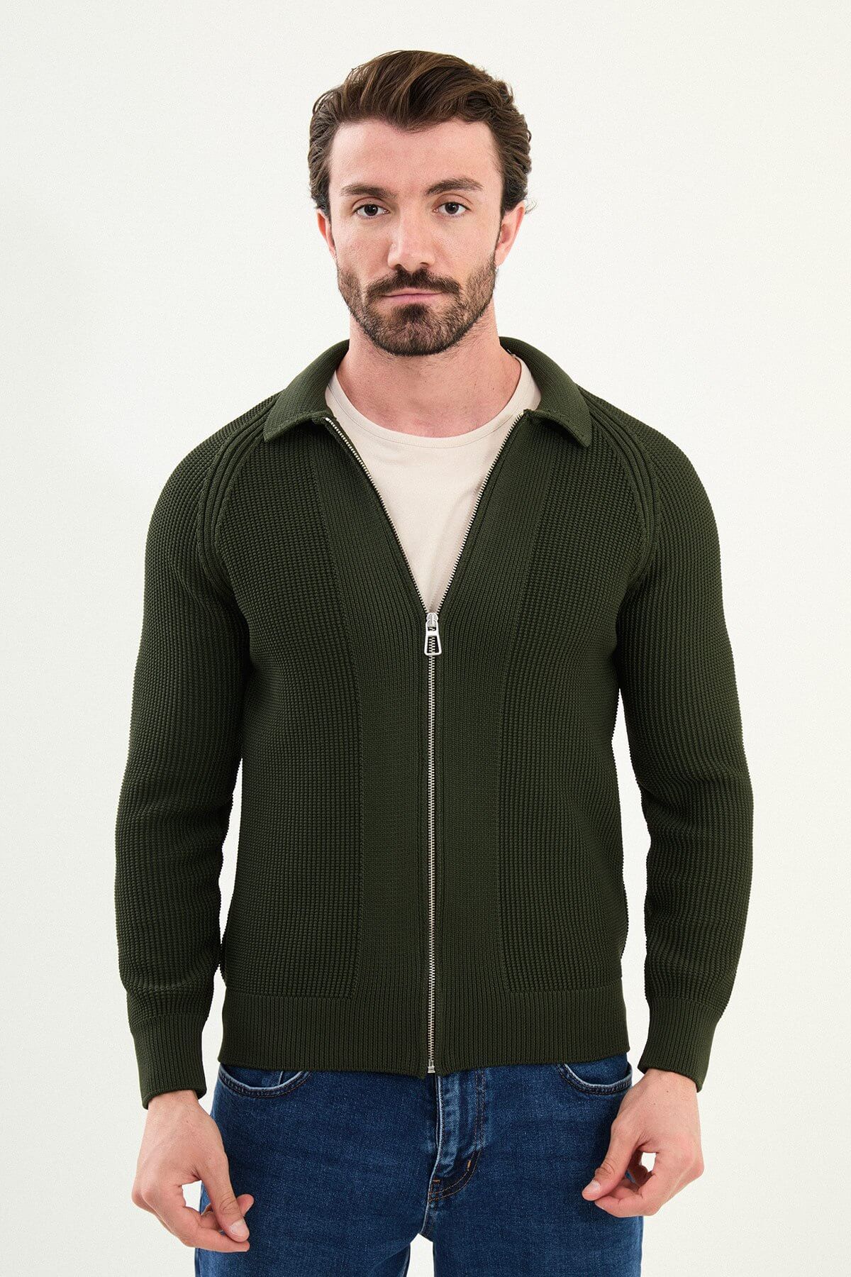 A man wearing a slim fit green ribbed zip-up knitwear cardigan with a collared design, paired with blue jeans and beige sneakers.