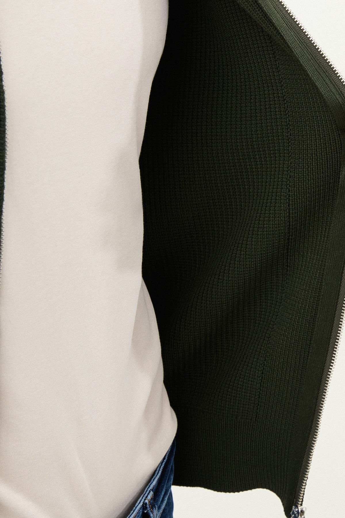 A man wearing a slim fit green ribbed zip-up knitwear cardigan with a collared design, paired with blue jeans and beige sneakers.