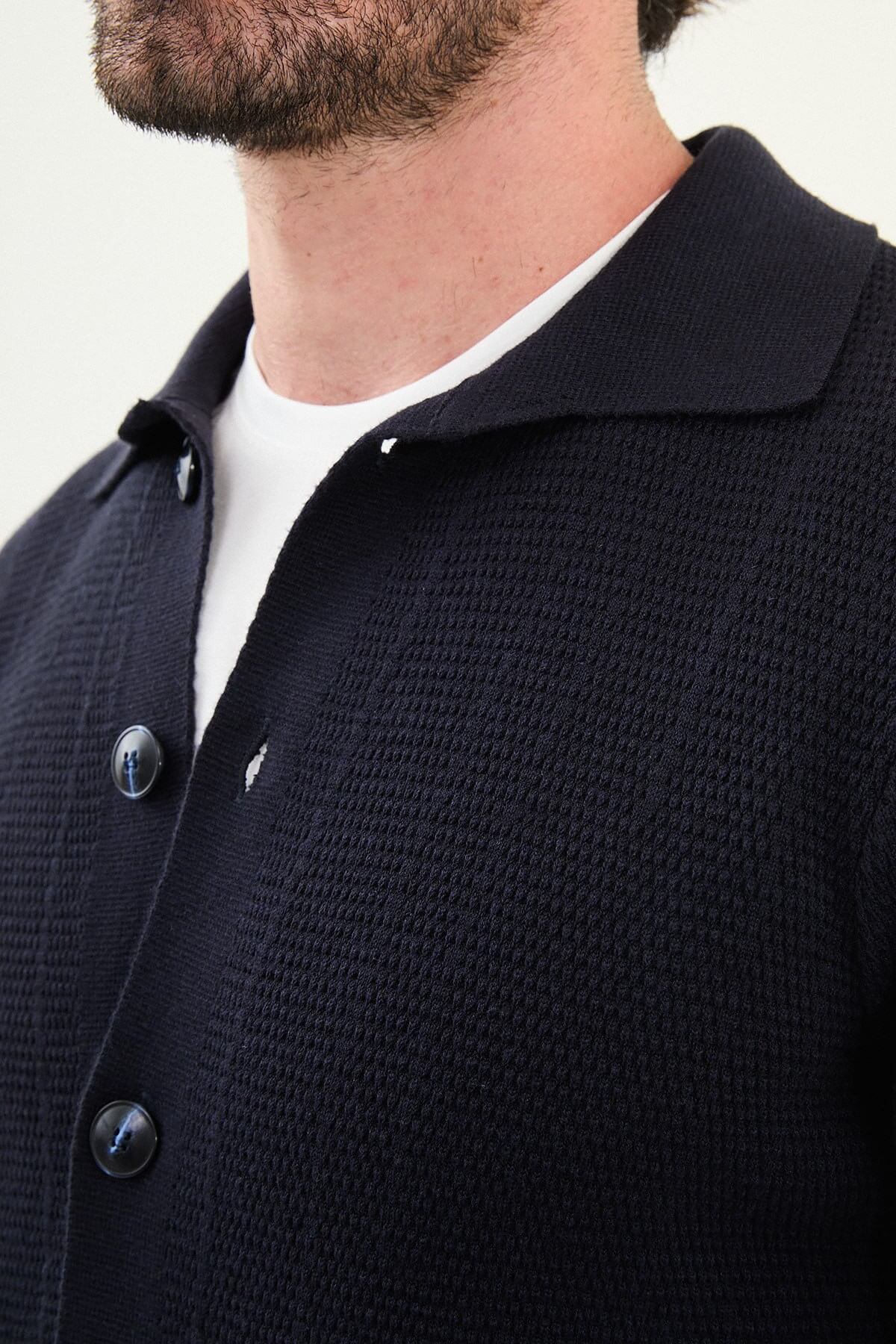 A man wearing a slim fit navy blue button-up cardigan with front pockets, styled with a white T-shirt and blue jeans.