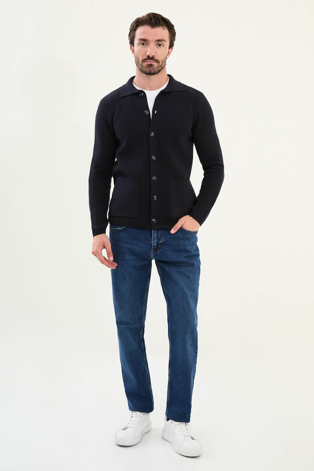A man wearing a slim fit navy blue button-up cardigan with front pockets, styled with a white T-shirt and blue jeans.