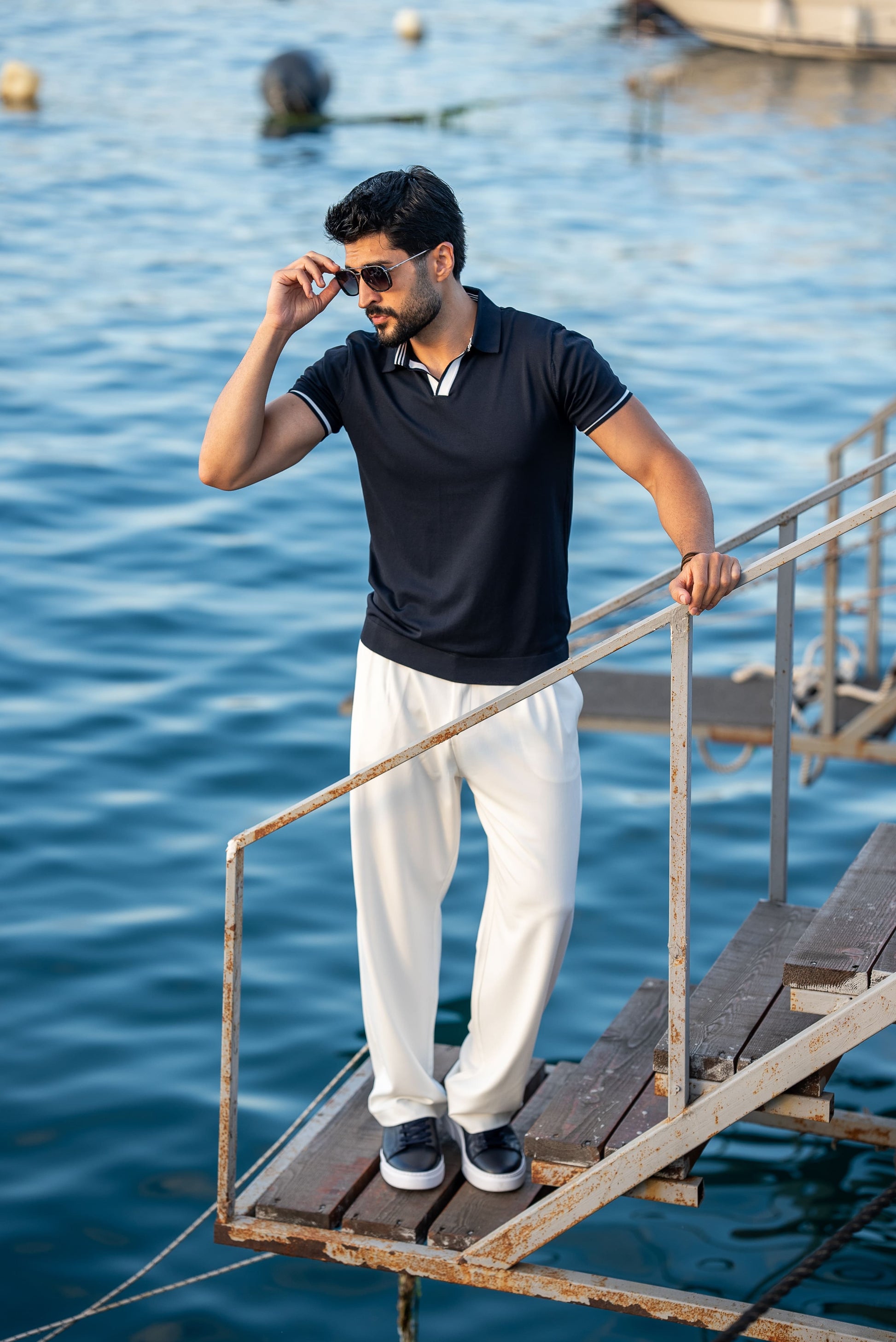 A stylish man in a navy blue polo shirt with contrast tipping and white flowing trousers, standing on a rustic dock above calm blue water on a sunny summer day.