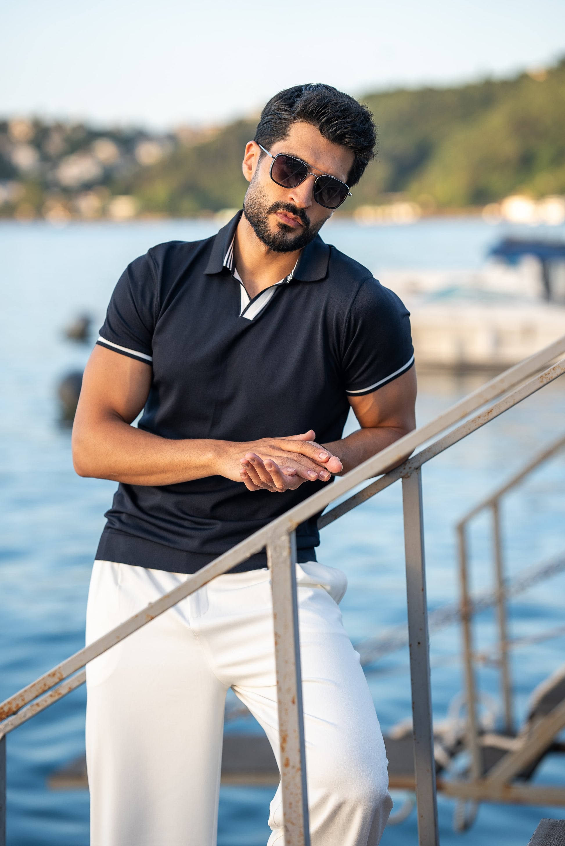 A stylish man in a navy blue polo shirt with contrast tipping and white flowing trousers, standing on a rustic dock above calm blue water on a sunny summer day.