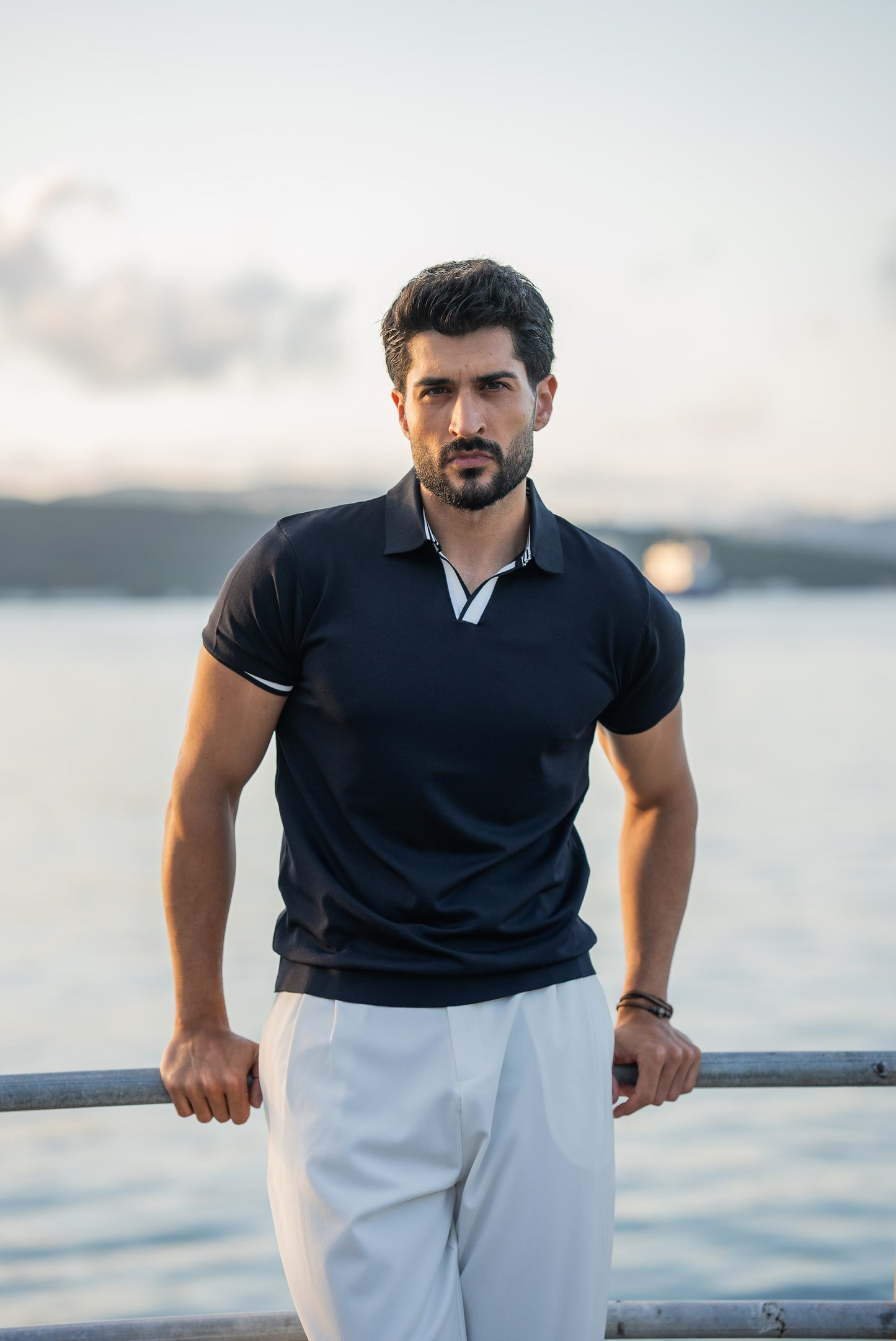 A stylish man in a navy blue polo shirt with contrast tipping and white flowing trousers, standing on a rustic dock above calm blue water on a sunny summer day.