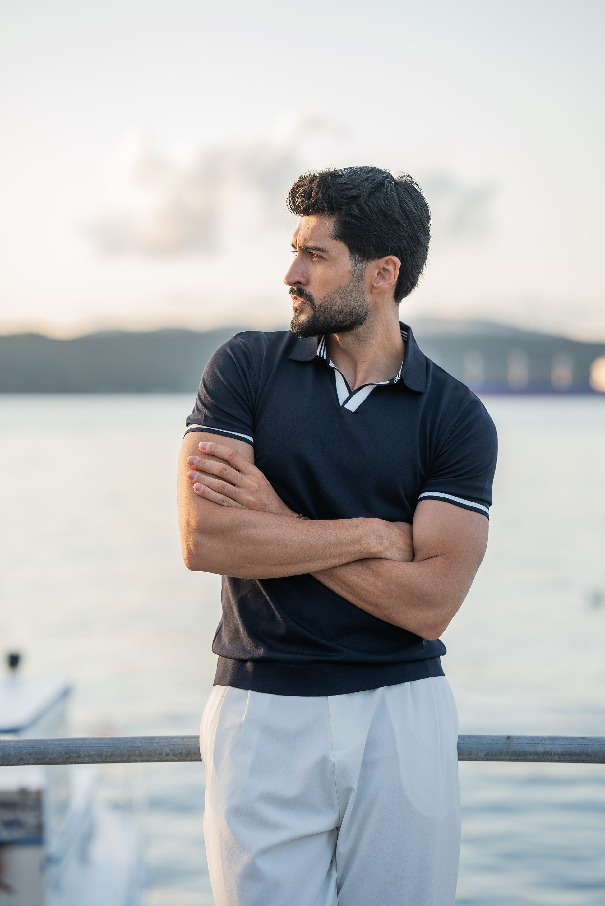 A stylish man in a navy blue polo shirt with contrast tipping and white flowing trousers, standing on a rustic dock above calm blue water on a sunny summer day.