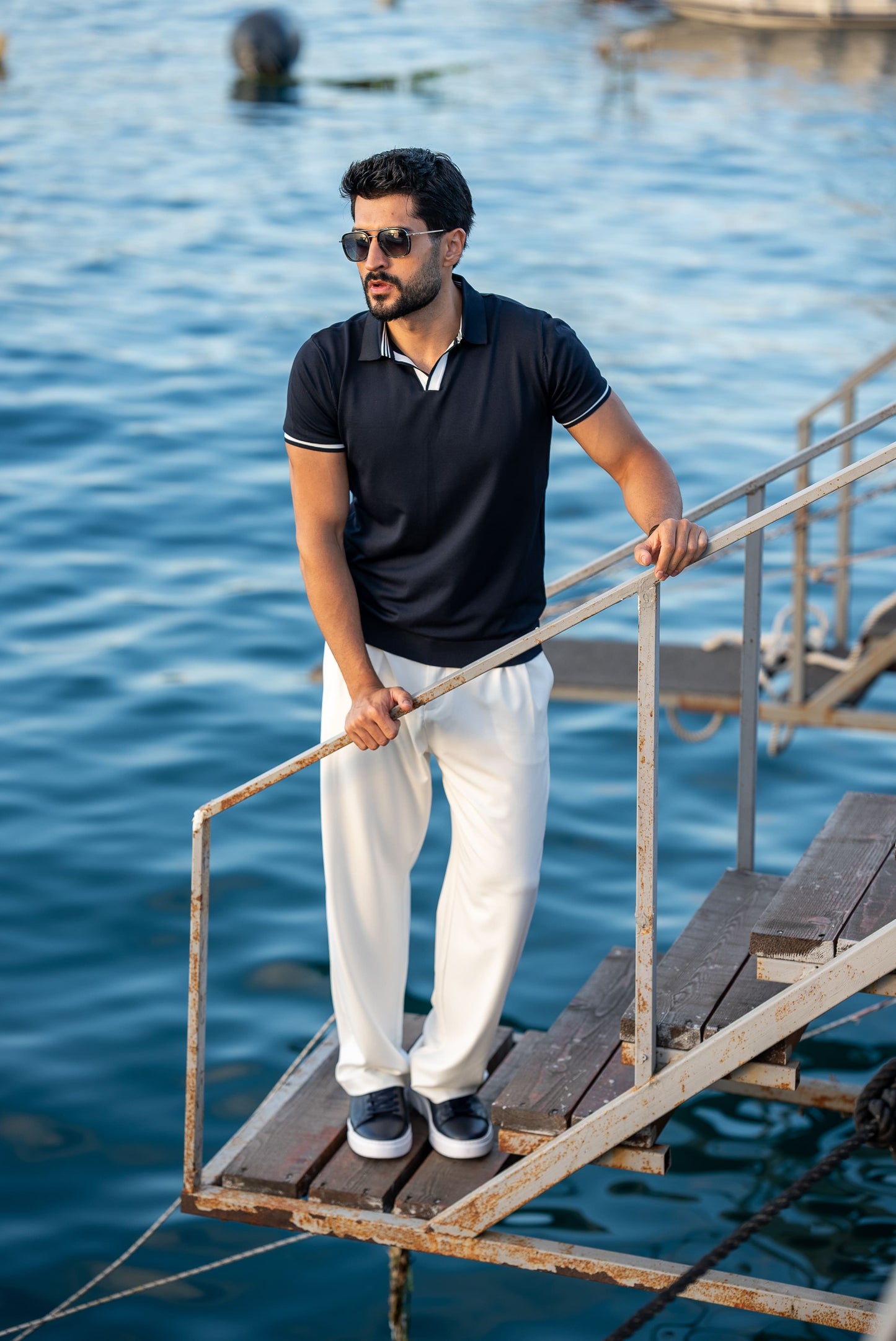 A stylish man in a navy blue polo shirt with contrast tipping and white flowing trousers, standing on a rustic dock above calm blue water on a sunny summer day.