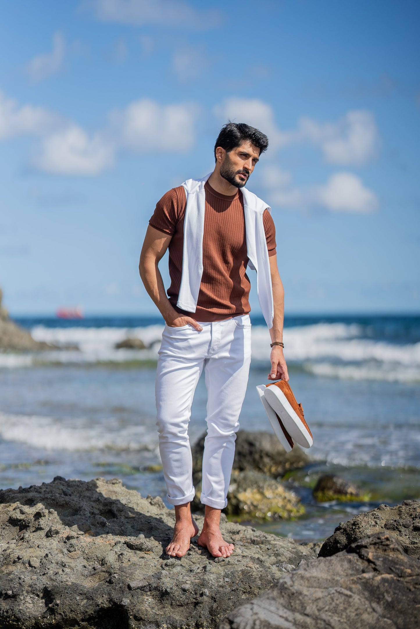 A man poses by the ocean in a ribbed tile-brown short sleeve knitted t-shirt paired with slim-fit white jeans. He has a white sweater draped over his shoulders, styled casually for a summer coastal vibe under a clear blue sky.