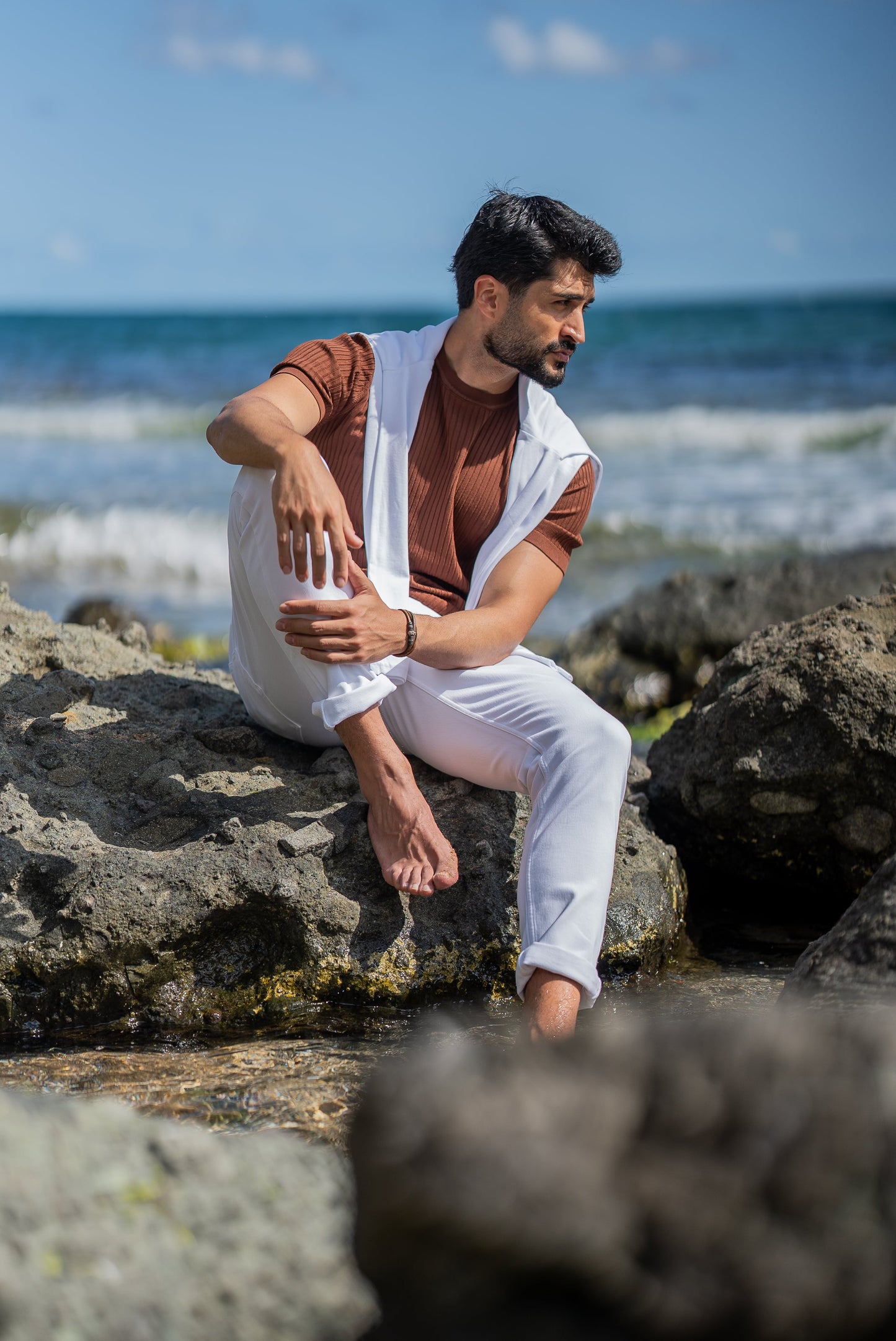 A man poses by the ocean in a ribbed tile-brown short sleeve knitted t-shirt paired with slim-fit white jeans. He has a white sweater draped over his shoulders, styled casually for a summer coastal vibe under a clear blue sky.