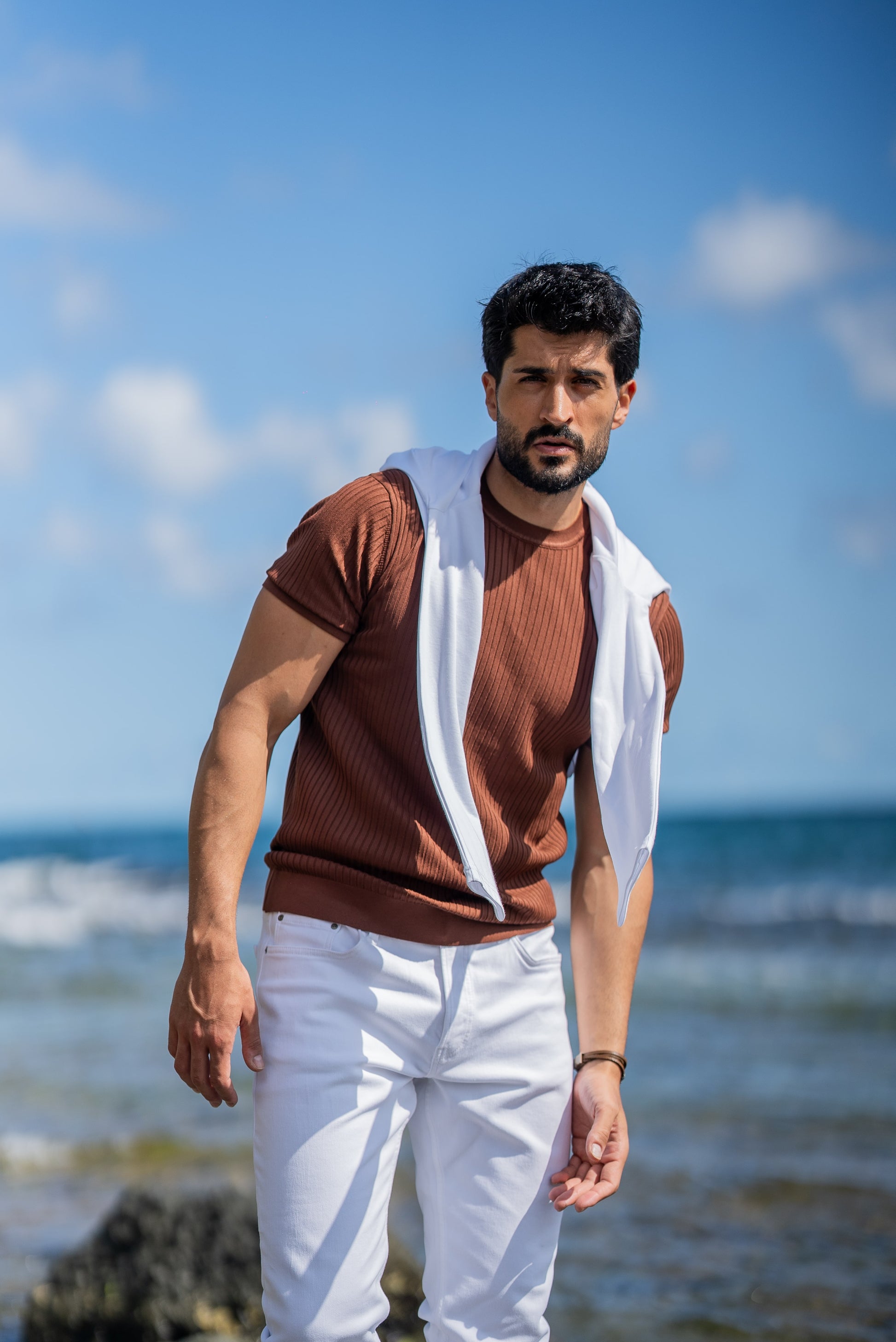 A man poses by the ocean in a ribbed tile-brown short sleeve knitted t-shirt paired with slim-fit white jeans. He has a white sweater draped over his shoulders, styled casually for a summer coastal vibe under a clear blue sky.