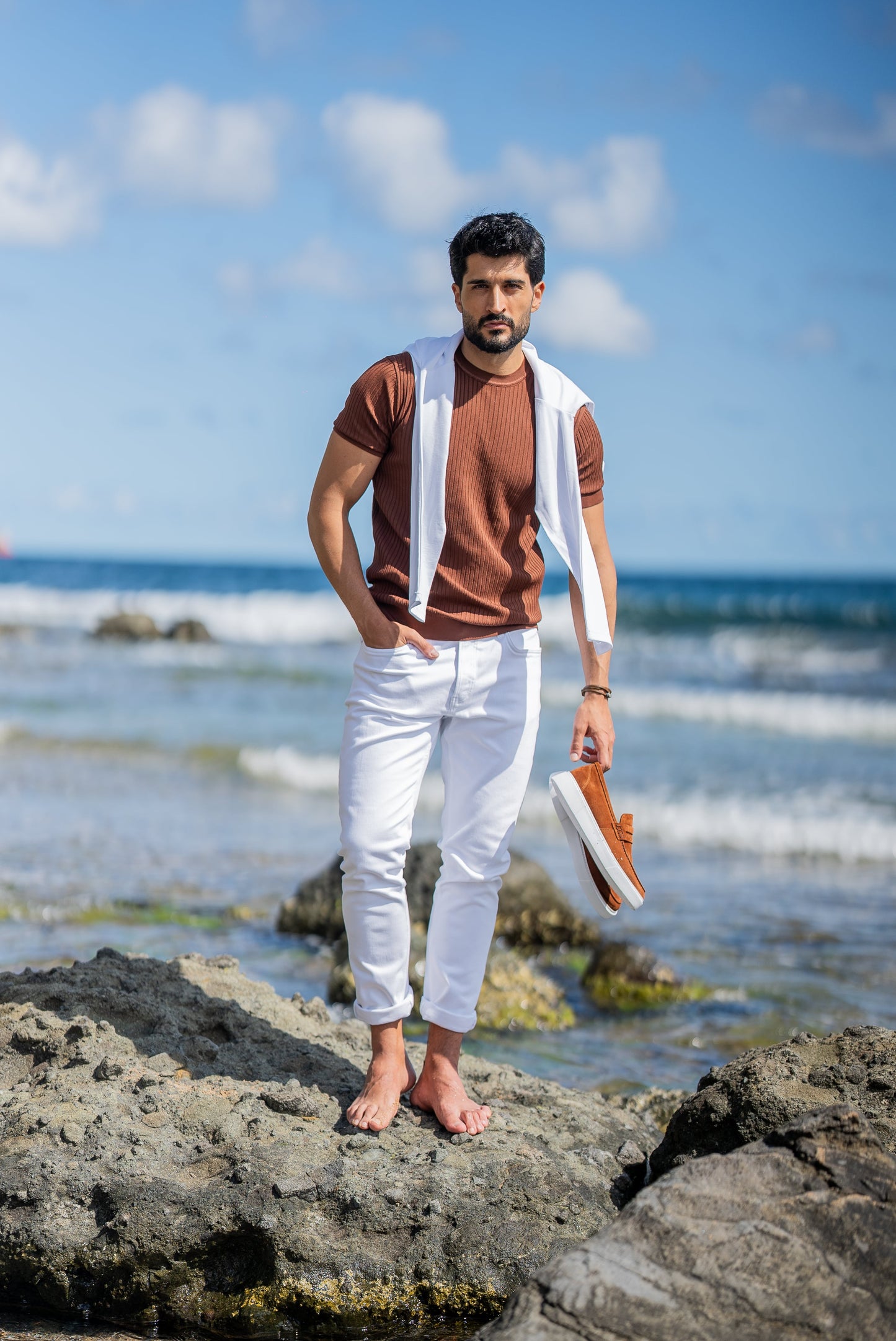 A man poses by the ocean in a ribbed tile-brown short sleeve knitted t-shirt paired with slim-fit white jeans. He has a white sweater draped over his shoulders, styled casually for a summer coastal vibe under a clear blue sky.