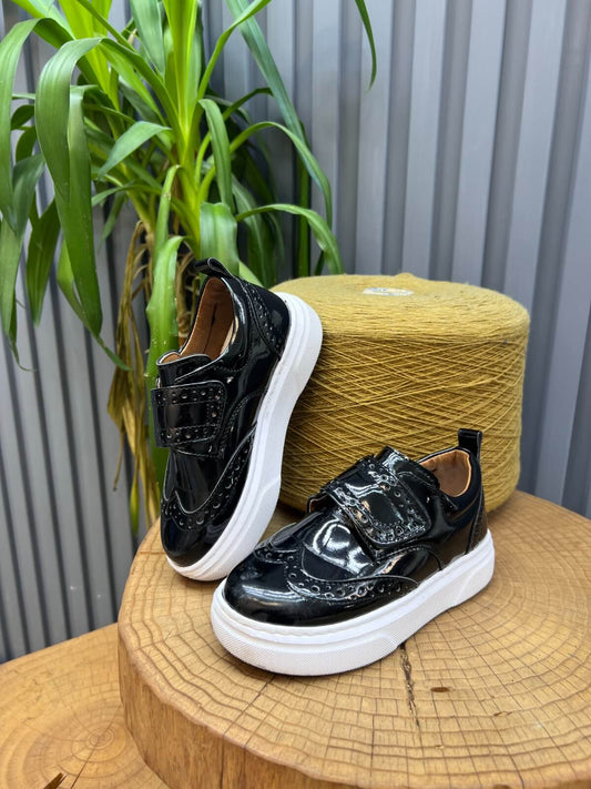 A pair of glossy black leather kids’ brogue slip-on shoes with white EVA soles and Velcro straps, displayed on a wooden platform beside green plants.