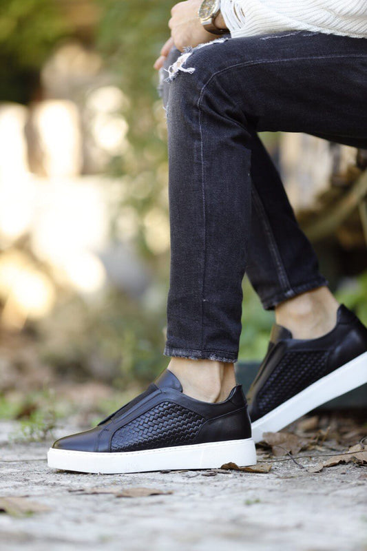 Black leather slip-on sneakers with woven side panels and white sole, styled with black jeans and cream sweater