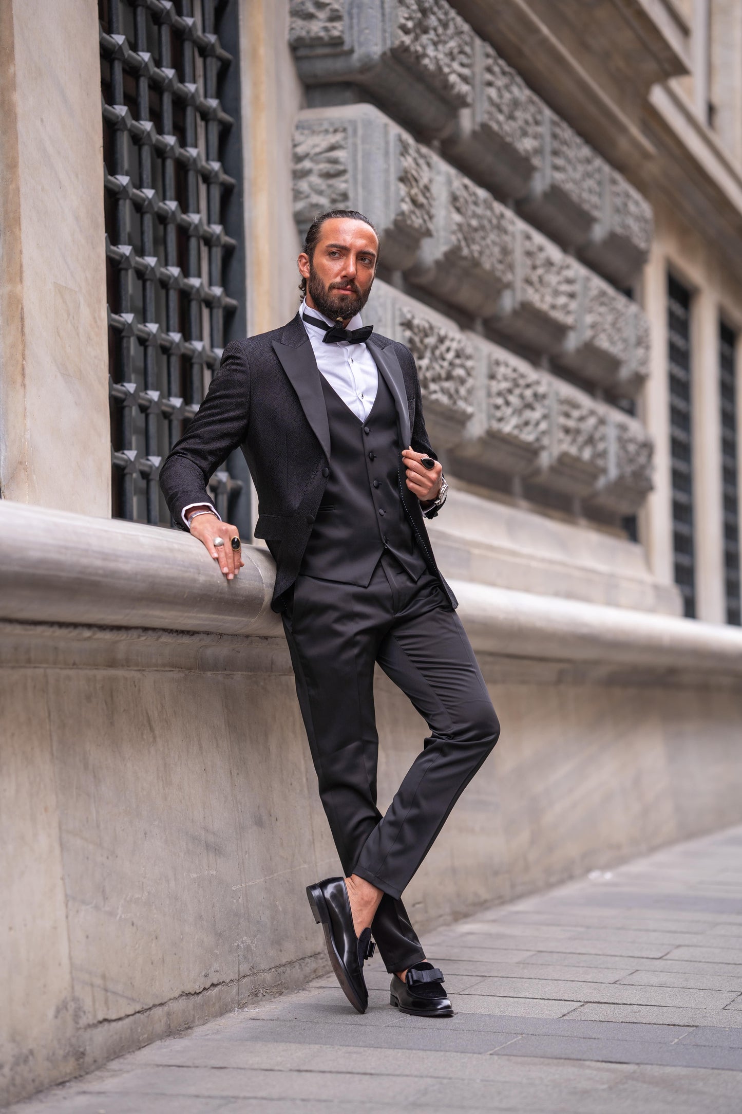 Slim-fit black tuxedo with double-button closure and pointed collar, modeled for fall-winter weddings and formal events — styled with matching vest, shirt, tie, and tailored trousers.