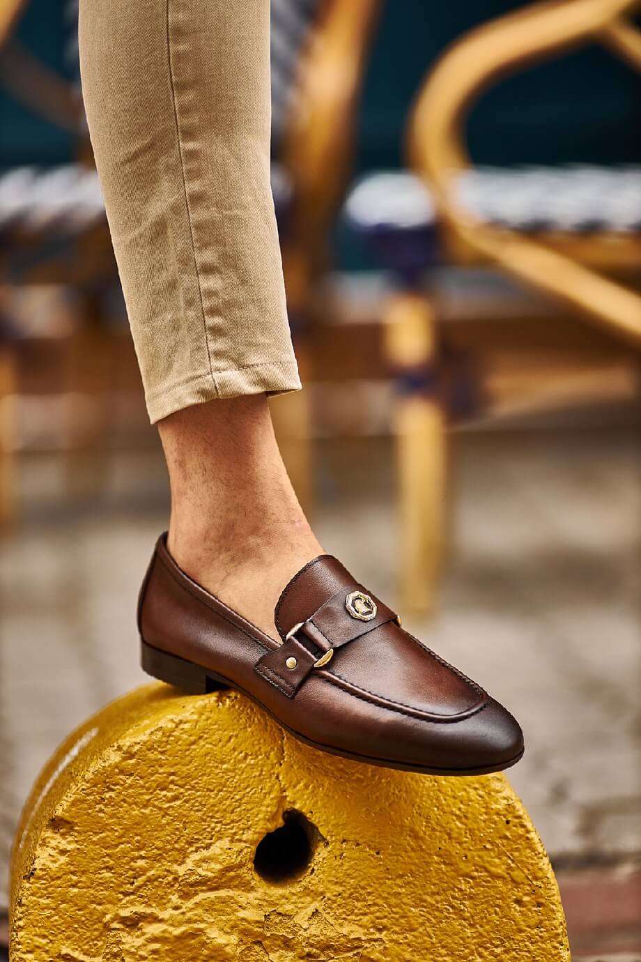 Brown leather loafers with gold buckle detail worn with beige pants, styled in a casual street-side café setting.