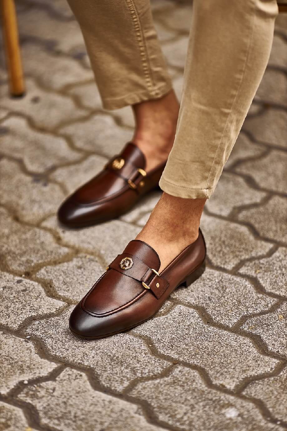 Brown leather loafers with gold buckle detail worn with beige pants, styled in a casual street-side café setting.