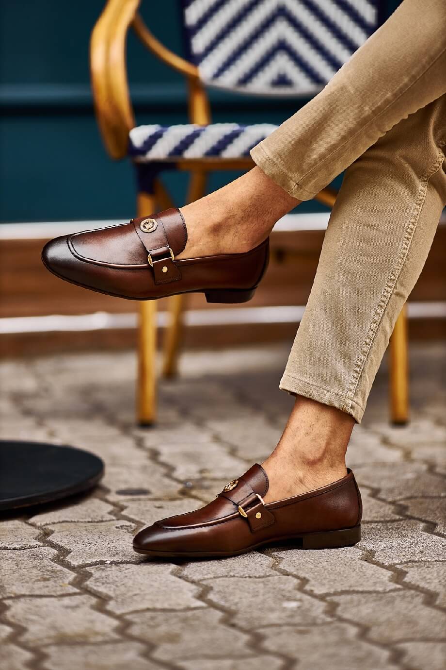 Brown leather loafers with gold buckle detail worn with beige pants, styled in a casual street-side café setting.
