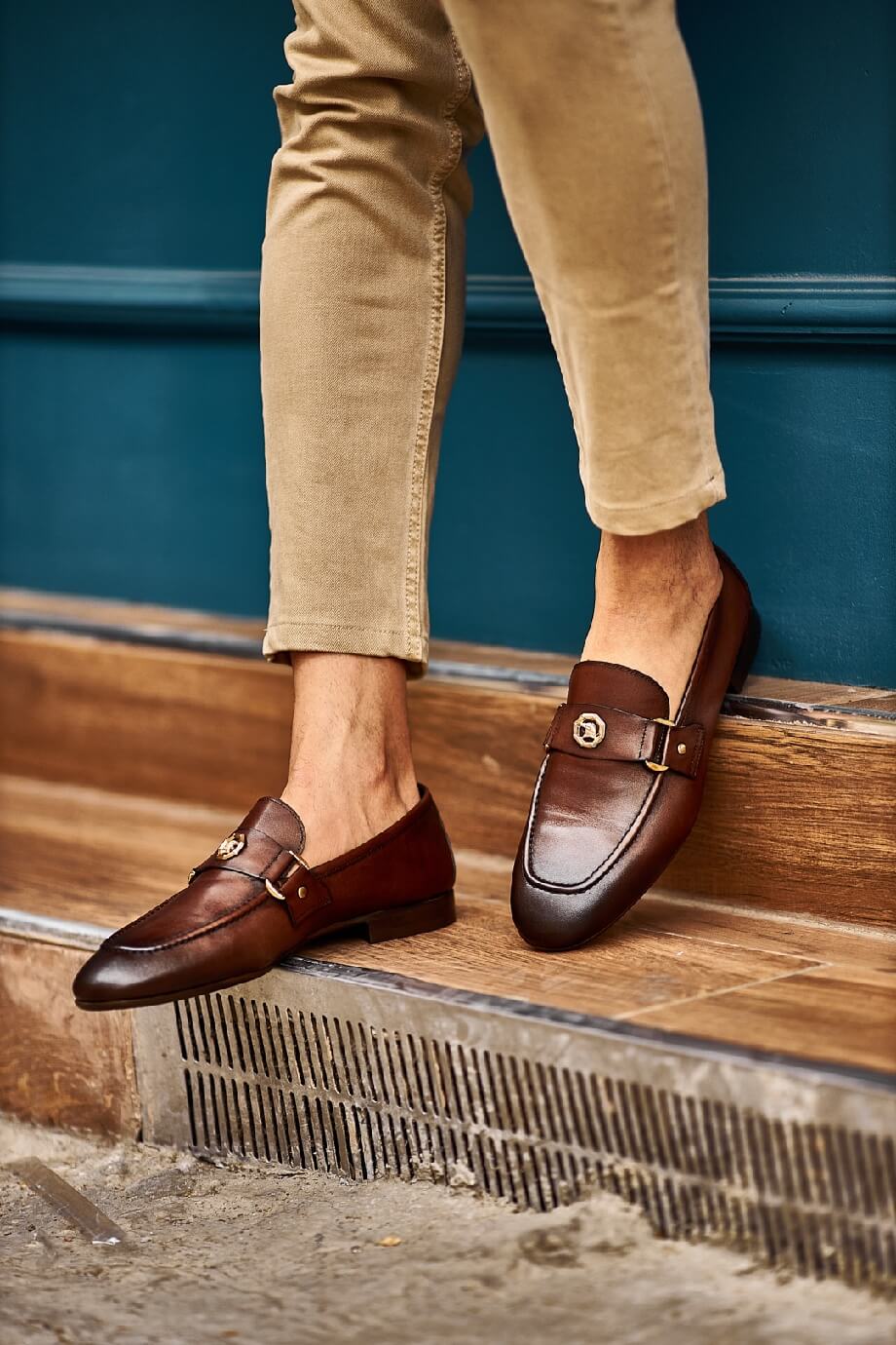 Brown leather loafers with gold buckle detail worn with beige pants, styled in a casual street-side café setting.
