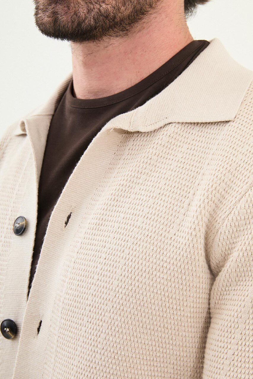A man wearing a slim fit beige button-up knit cardigan over a dark brown T-shirt, styled with dark brown corduroy trousers.