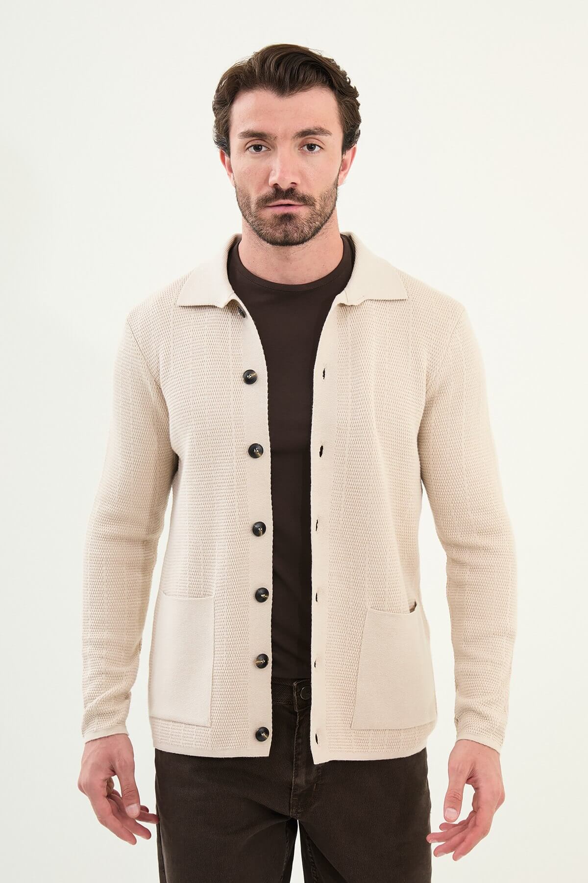 A man wearing a slim fit beige button-up knit cardigan over a dark brown T-shirt, styled with dark brown corduroy trousers.