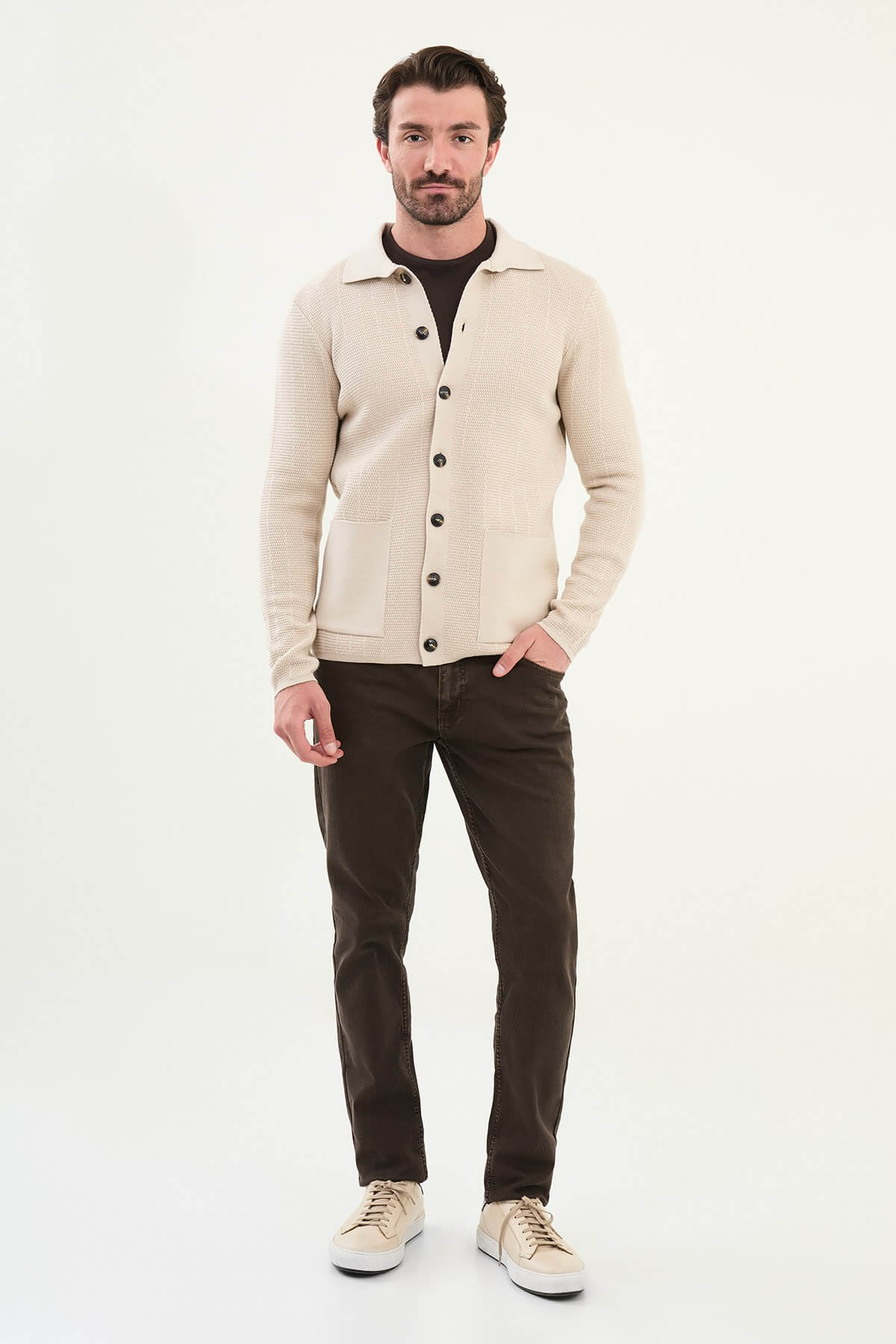 A man wearing a slim fit beige button-up knit cardigan over a dark brown T-shirt, styled with dark brown corduroy trousers.