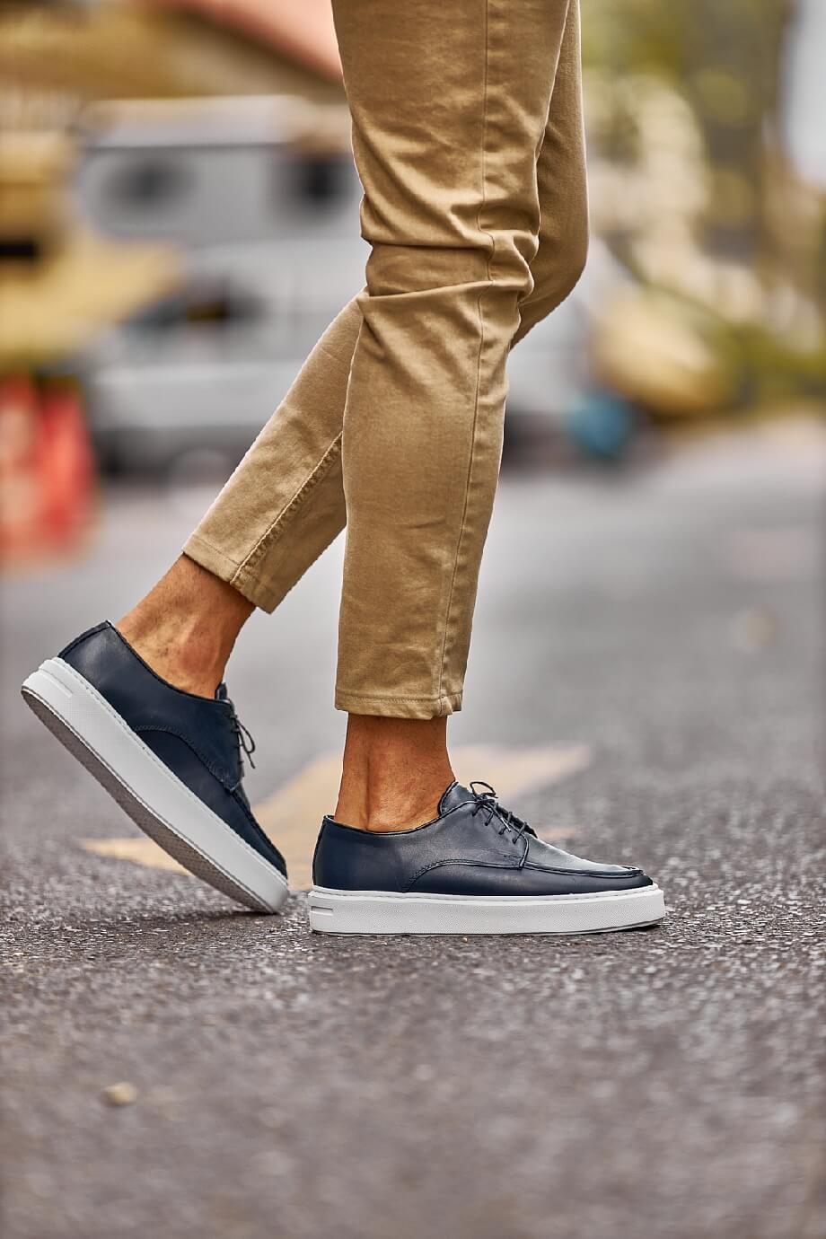 Close-up of a man walking in Tulum navy leather lace-up derby shoes with white EVA soles, styled with cropped khaki trousers on a sunlit street.