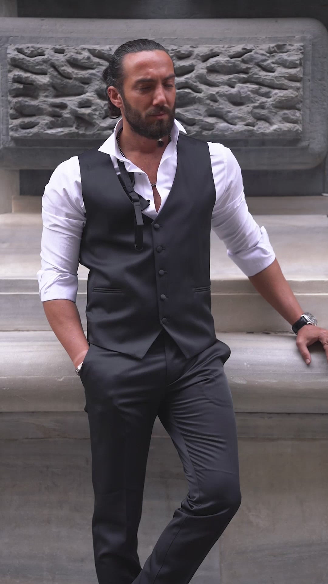 Slim-fit black tuxedo with double-button closure and pointed collar, modeled for fall-winter weddings and formal events — styled with matching vest, shirt, tie, and tailored trousers.