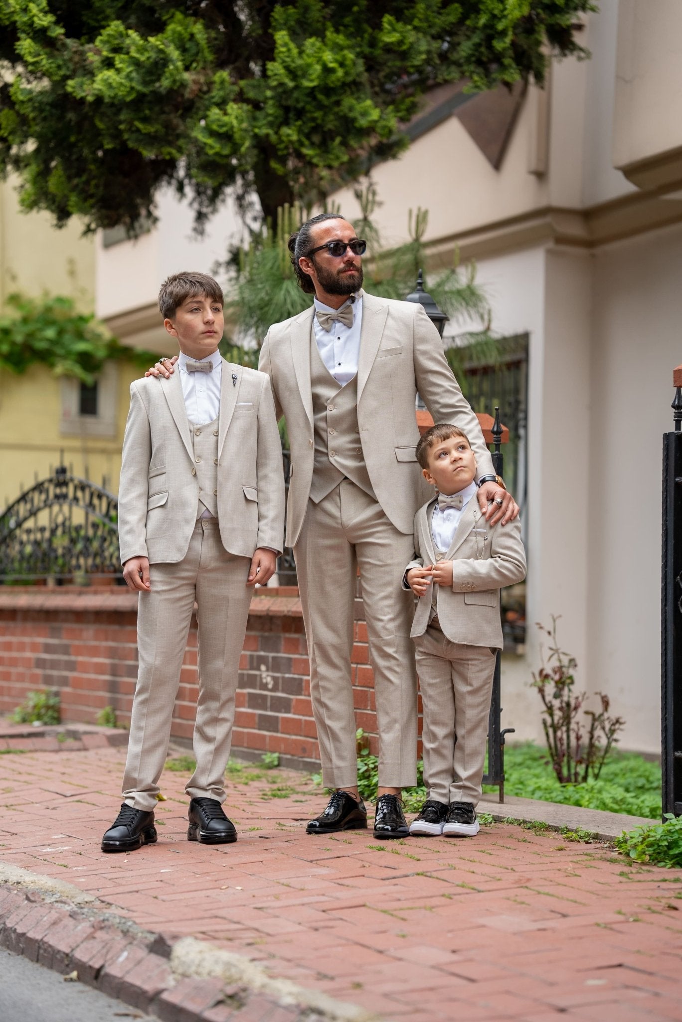 Father and his two sons walking down a street in matching beige tuxedos with elegant bow ties, exuding confidence and style during a formal occasion.