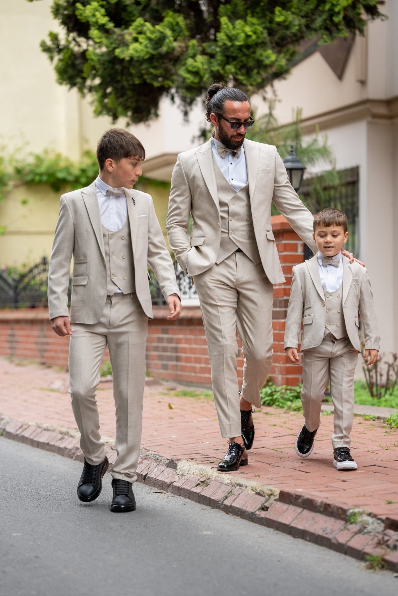 Father and his two sons walking down a street in matching beige tuxedos with elegant bow ties, exuding confidence and style during a formal occasion.