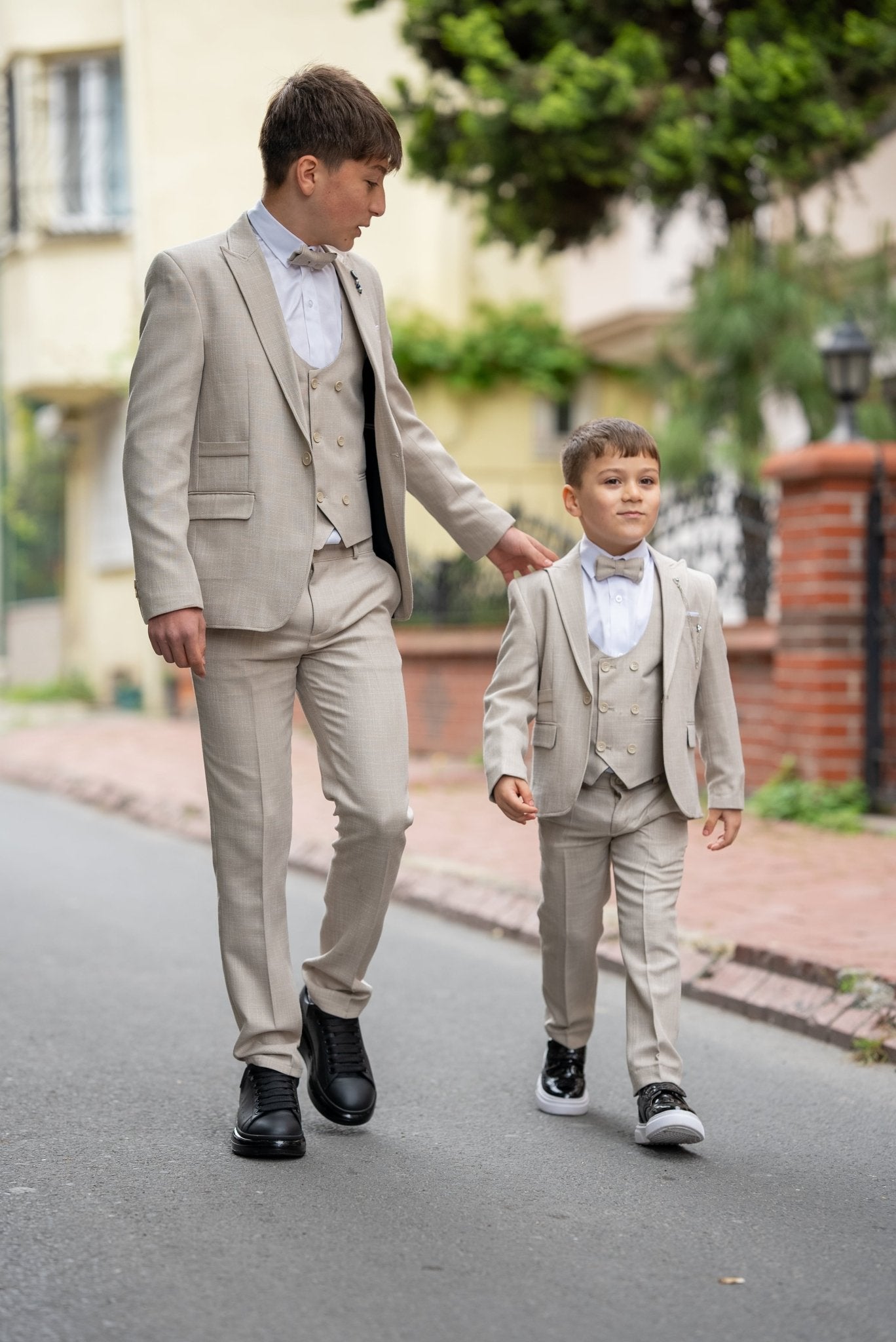 Father and his two sons walking down a street in matching beige tuxedos with elegant bow ties, exuding confidence and style during a formal occasion.
