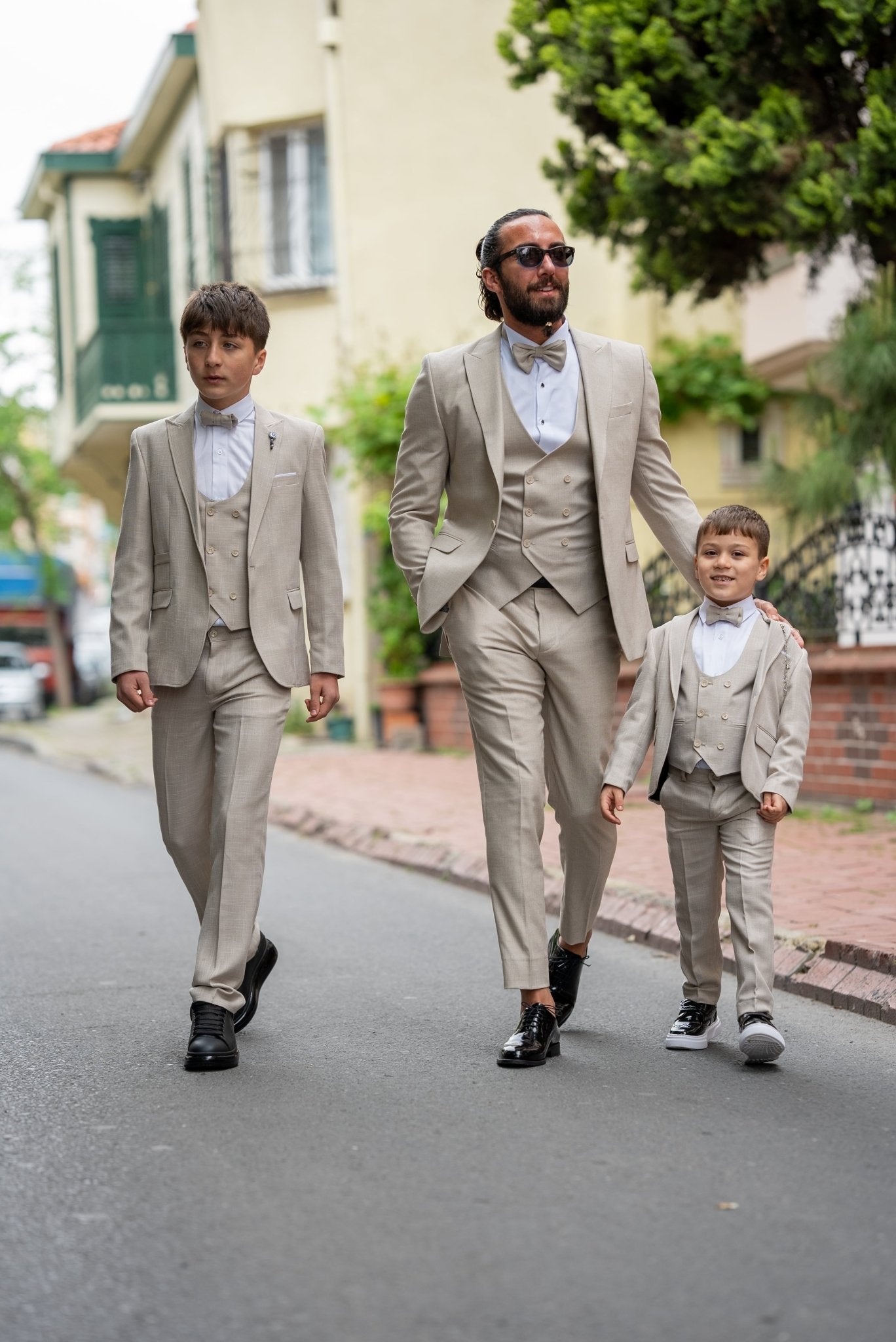 Father and his two sons walking down a street in matching beige tuxedos with elegant bow ties, exuding confidence and style during a formal occasion.