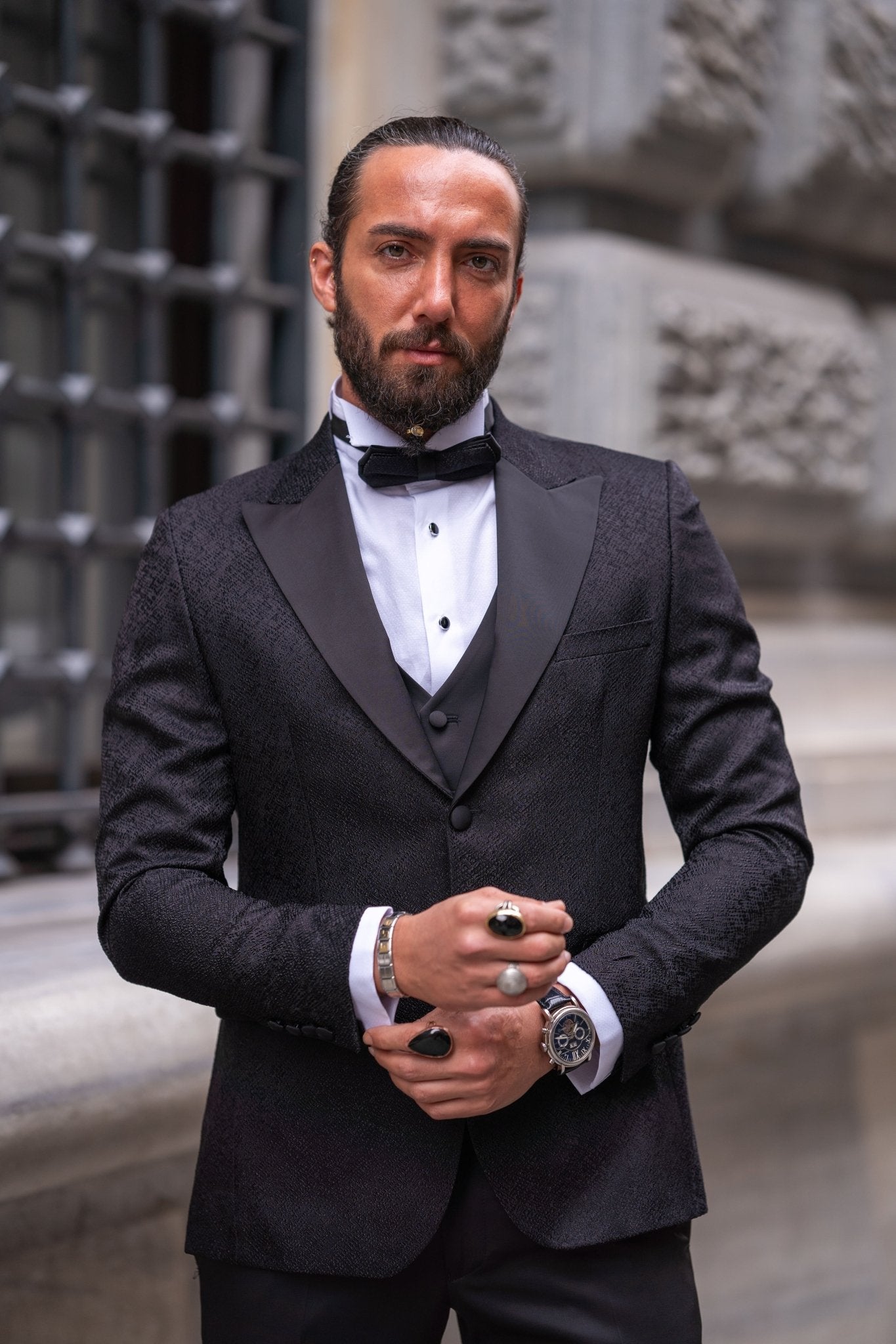 Slim-fit black tuxedo with double-button closure and pointed collar, modeled for fall-winter weddings and formal events — styled with matching vest, shirt, tie, and tailored trousers.