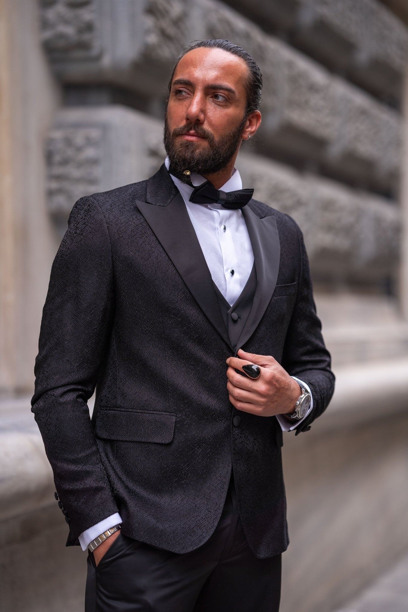 Slim-fit black tuxedo with double-button closure and pointed collar, modeled for fall-winter weddings and formal events — styled with matching vest, shirt, tie, and tailored trousers.