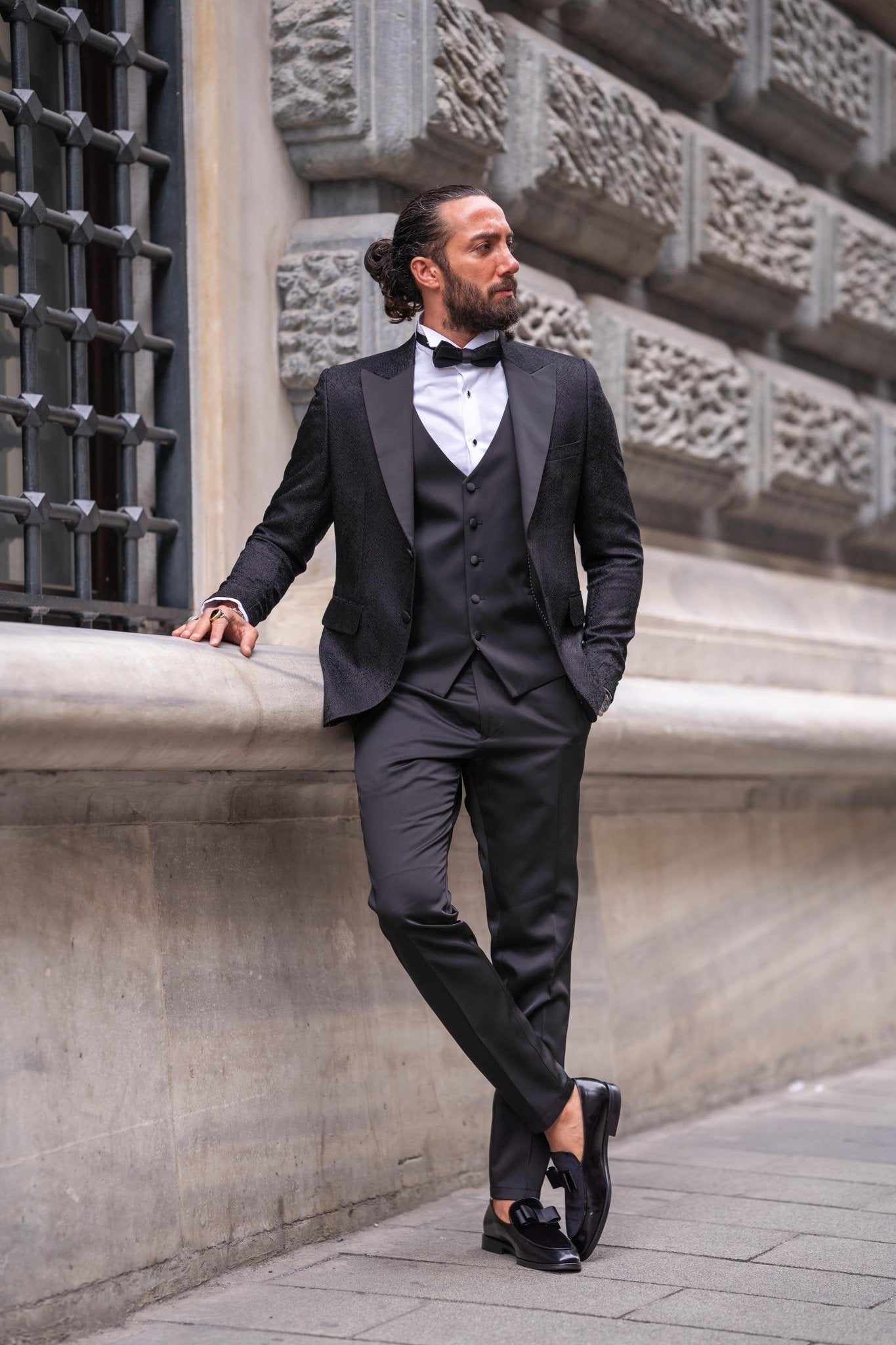 Slim-fit black tuxedo with double-button closure and pointed collar, modeled for fall-winter weddings and formal events — styled with matching vest, shirt, tie, and tailored trousers.