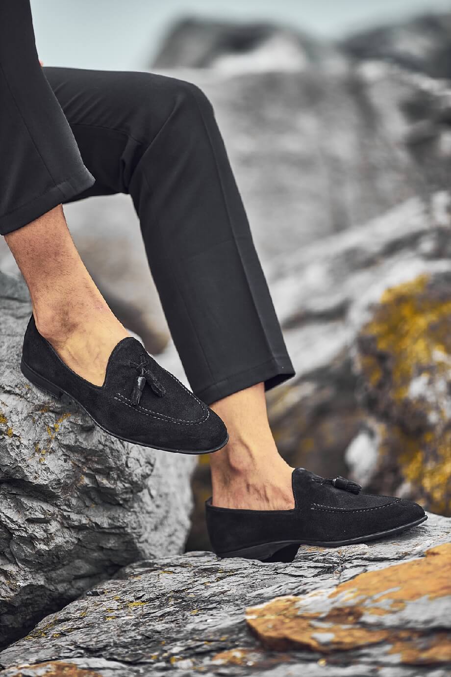 Man wearing black suede tassel loafers on rocky coastal terrain with cropped black pants, showcasing stylish summer footwear.