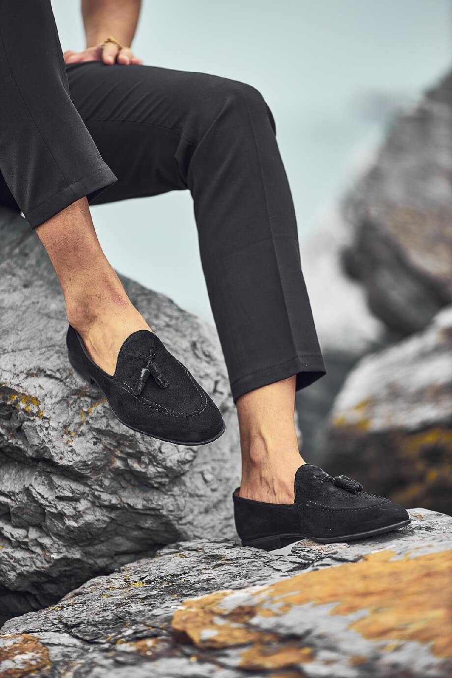 Man wearing black suede tassel loafers on rocky coastal terrain with cropped black pants, showcasing stylish summer footwear.
