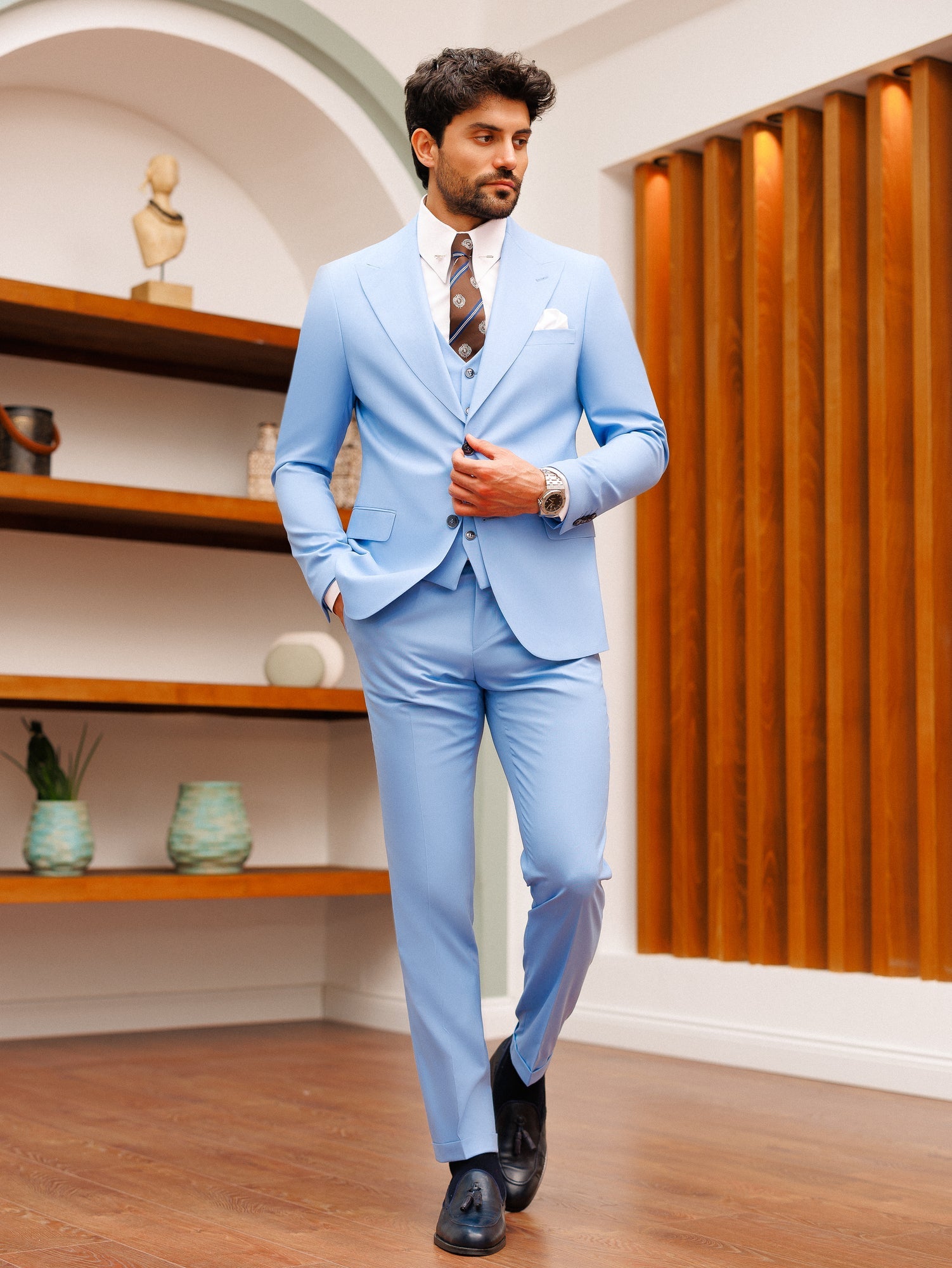 Men’s sky-blue slim-fit 3-piece suit with blazer, waistcoat, and trousers, styled with a white shirt and patterned tie in a modern interior setting.
