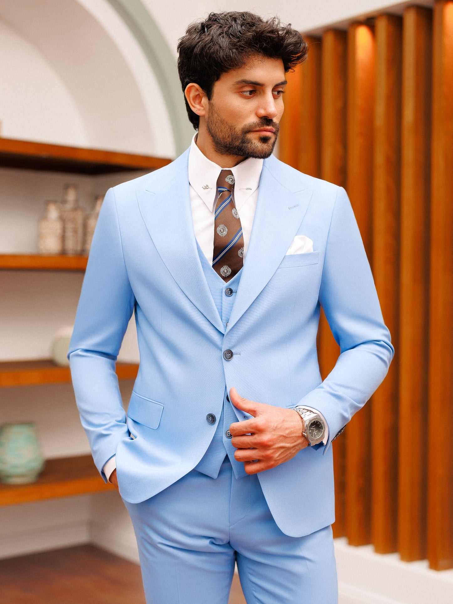 Men’s sky-blue slim-fit 3-piece suit with blazer, waistcoat, and trousers, styled with a white shirt and patterned tie in a modern interior setting.