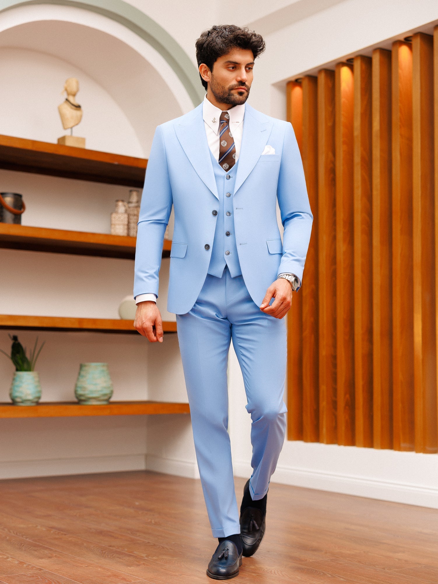 Men’s sky-blue slim-fit 3-piece suit with blazer, waistcoat, and trousers, styled with a white shirt and patterned tie in a modern interior setting.