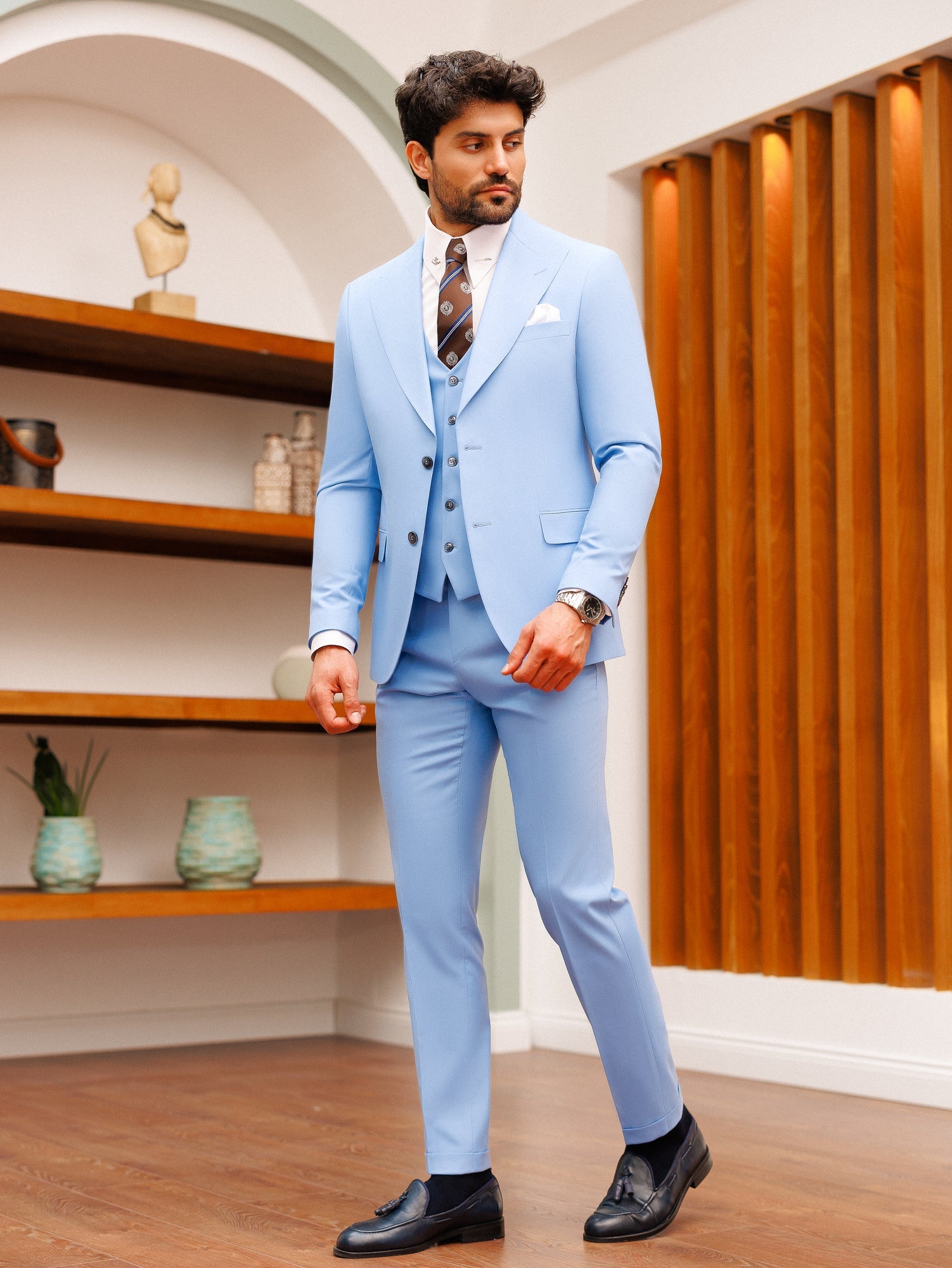 Men’s sky-blue slim-fit 3-piece suit with blazer, waistcoat, and trousers, styled with a white shirt and patterned tie in a modern interior setting.