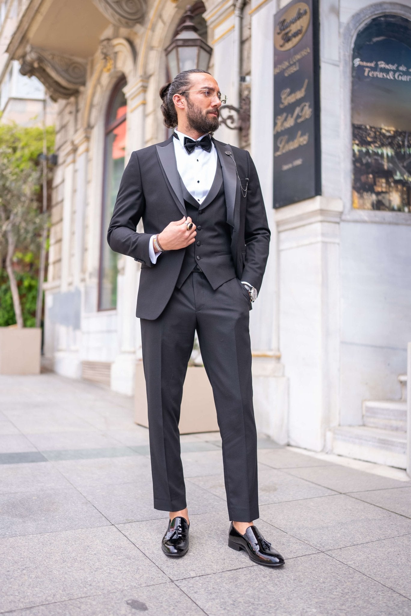 Black satin-collar men’s tuxedo in slim fit, styled with a vest, bow tie, and formal pants – a 5-piece formal set ideal for weddings, galas, and luxury events.