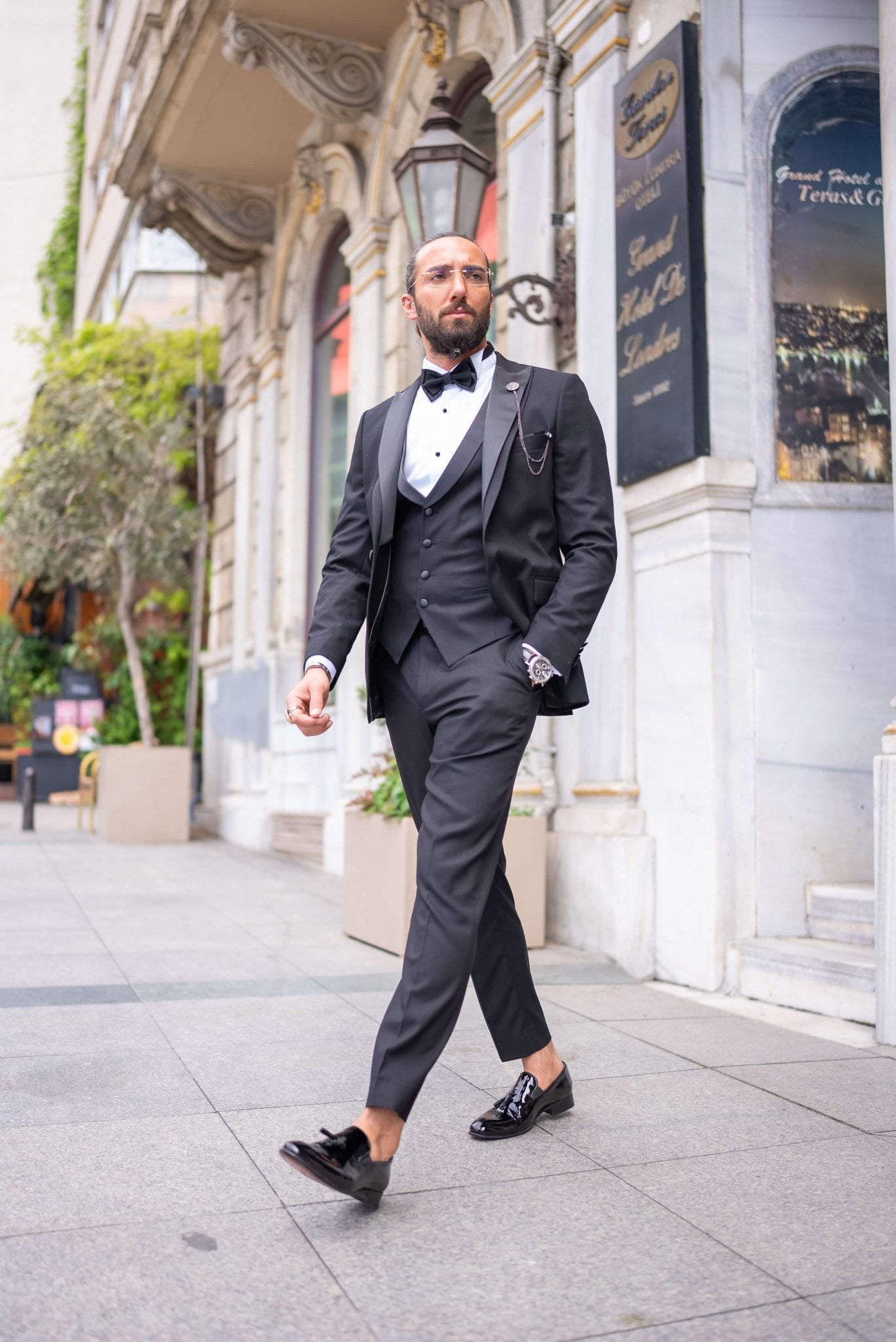 Black satin-collar men’s tuxedo in slim fit, styled with a vest, bow tie, and formal pants – a 5-piece formal set ideal for weddings, galas, and luxury events.