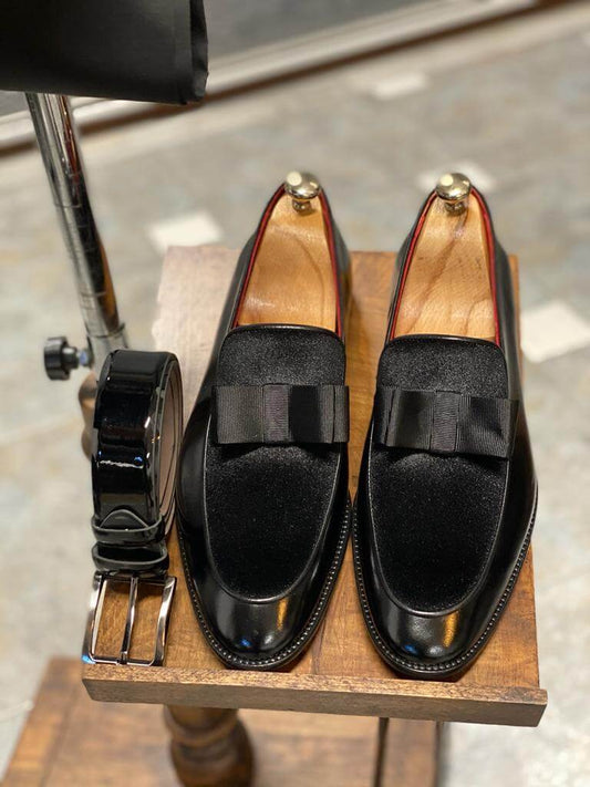 Elegant black tuxedo loafers with a sleek design and a stylish bow detail, placed on a light-colored carpet, accompanied by a matching black bow tie in a modern indoor setting.